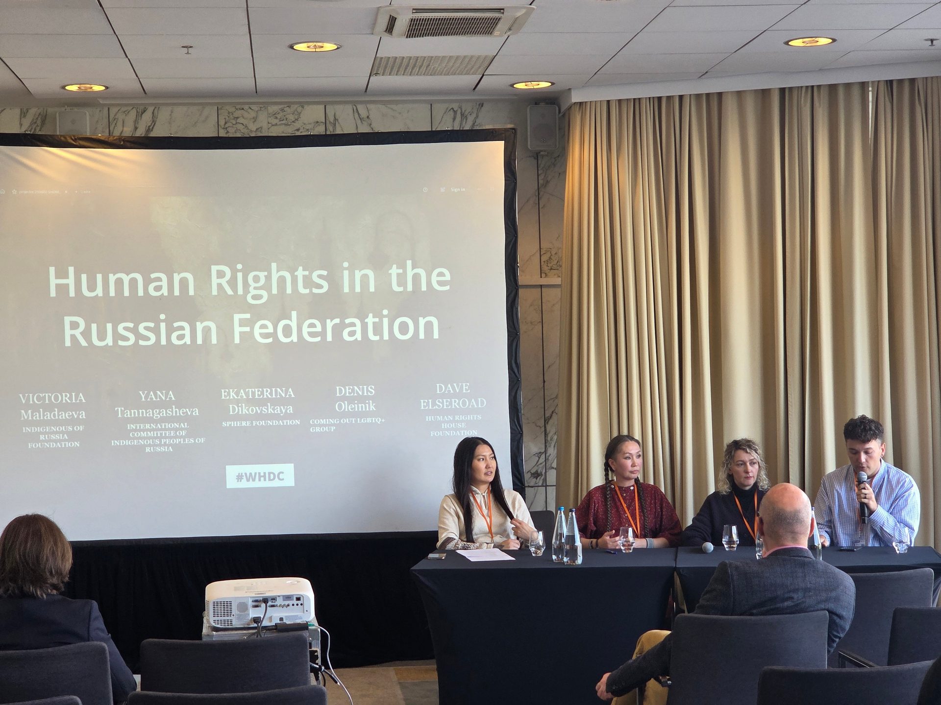 A panel discussion on 'Human Rights in the Russian Federation' with four speakers and a projection screen.
