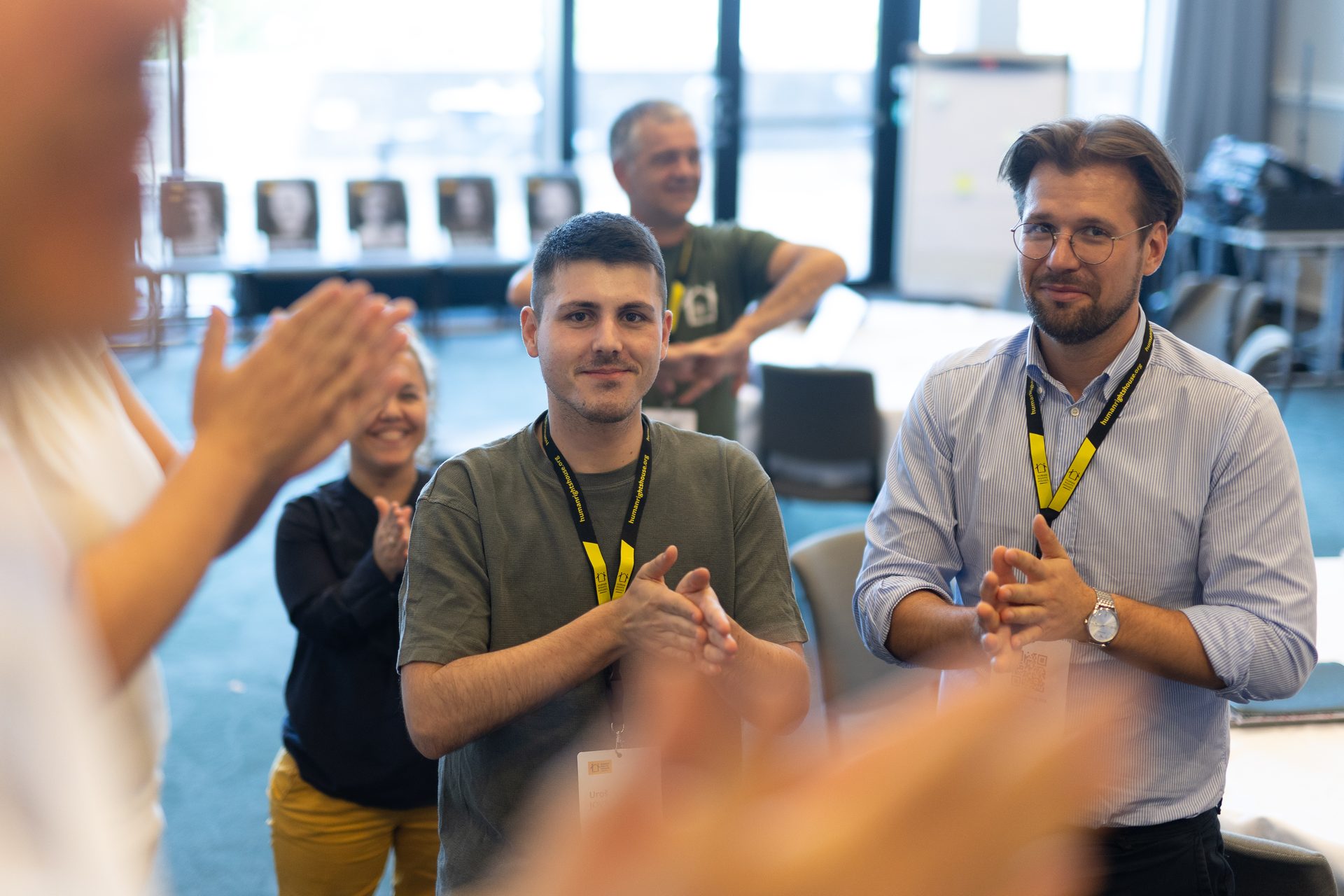 A group of people, including two men in the foreground, clap and smile during an indoor event.
