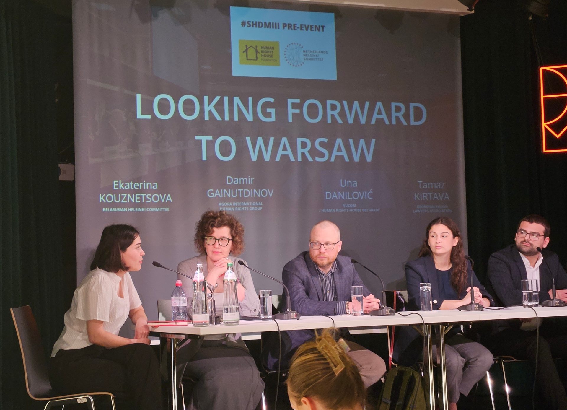 Five speakers at a panel discussion with a screen behind them, titled 'LOOKING FORWARD TO WARSAW'.
