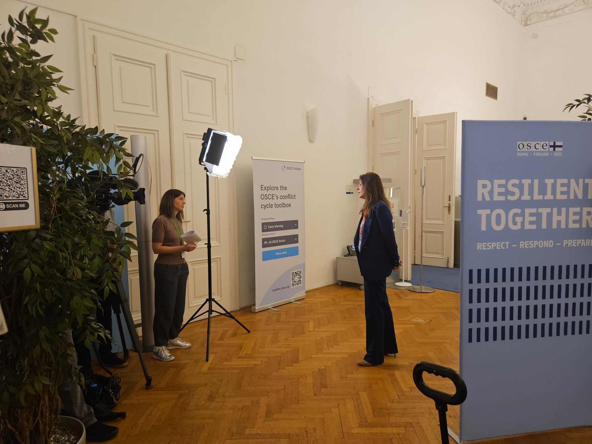 Two women, one speaking to the other, stand between OSCE banners and camera equipment in a large room.