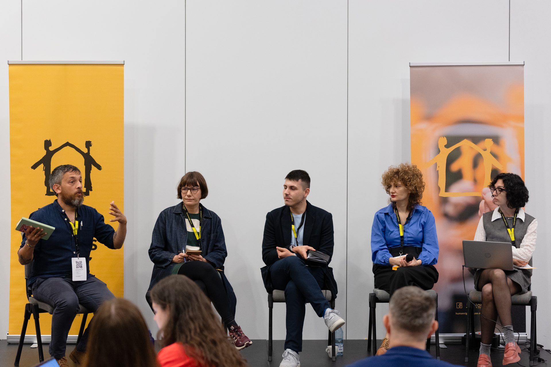 Five people sit on chairs on a panel discussion stage, with two yellow banners behind them.