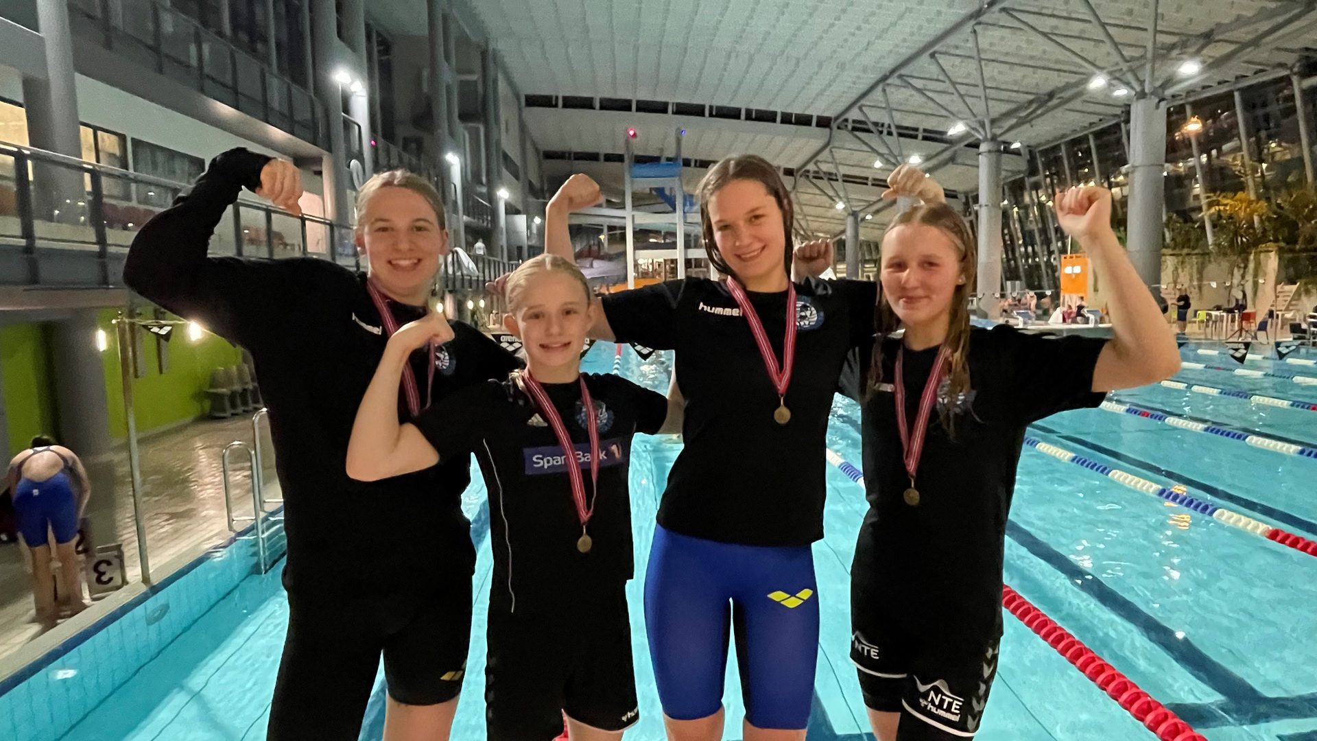 Four young female swimmers with medals proudly flex their biceps by a swimming pool.