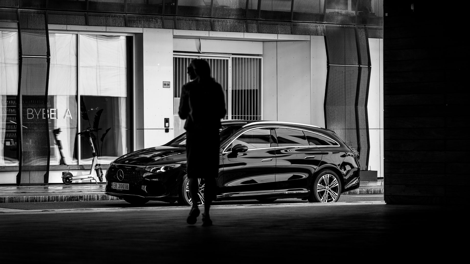 Black and white photo of a person walking past a parked black luxury station wagon in an urban setting.