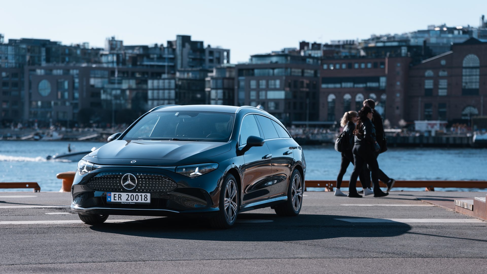 A black Mercedes-Benz EQS parked by a waterfront city, with people walking in the background.