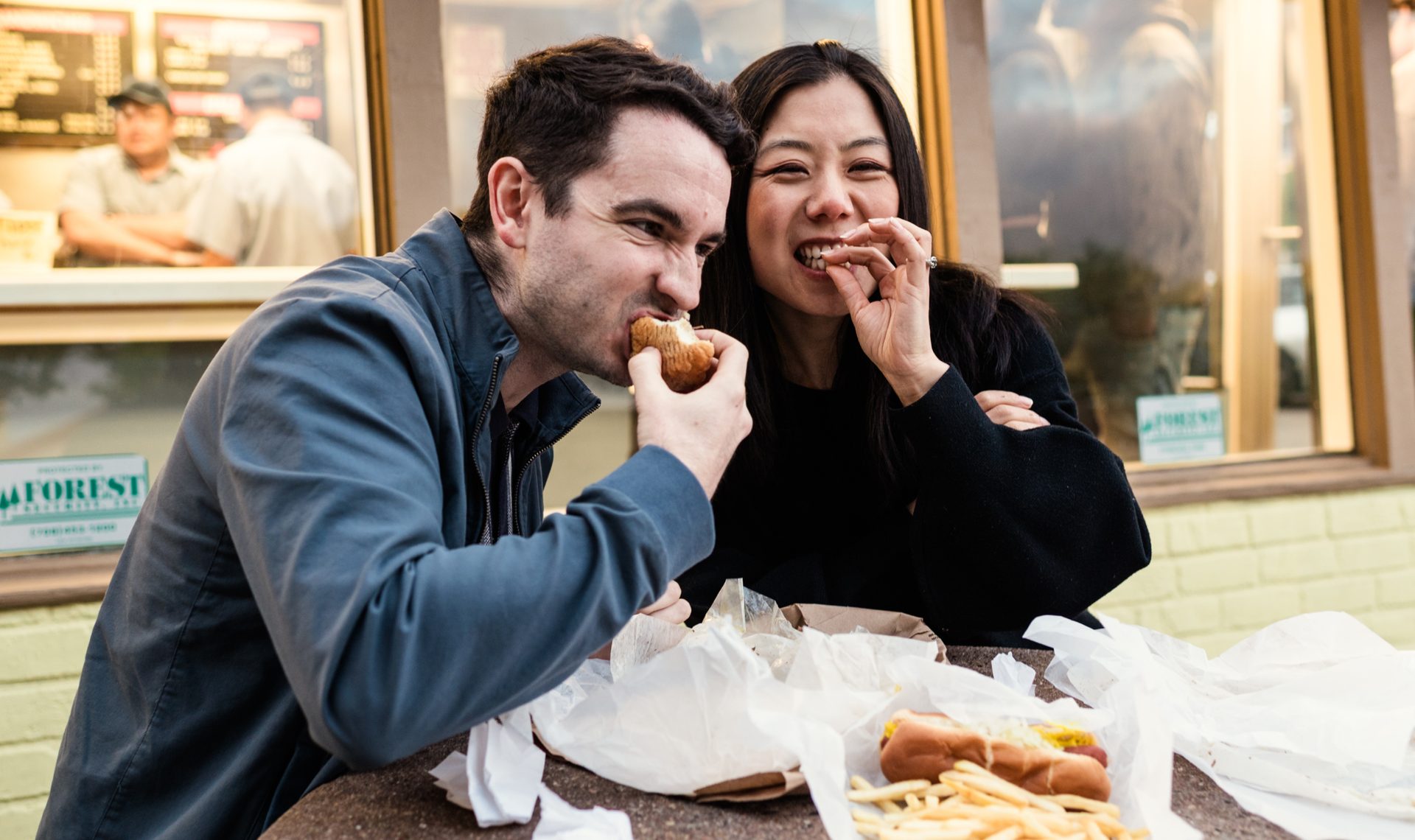 Man and woman smiling while eating hot dogs and fries outdoors.