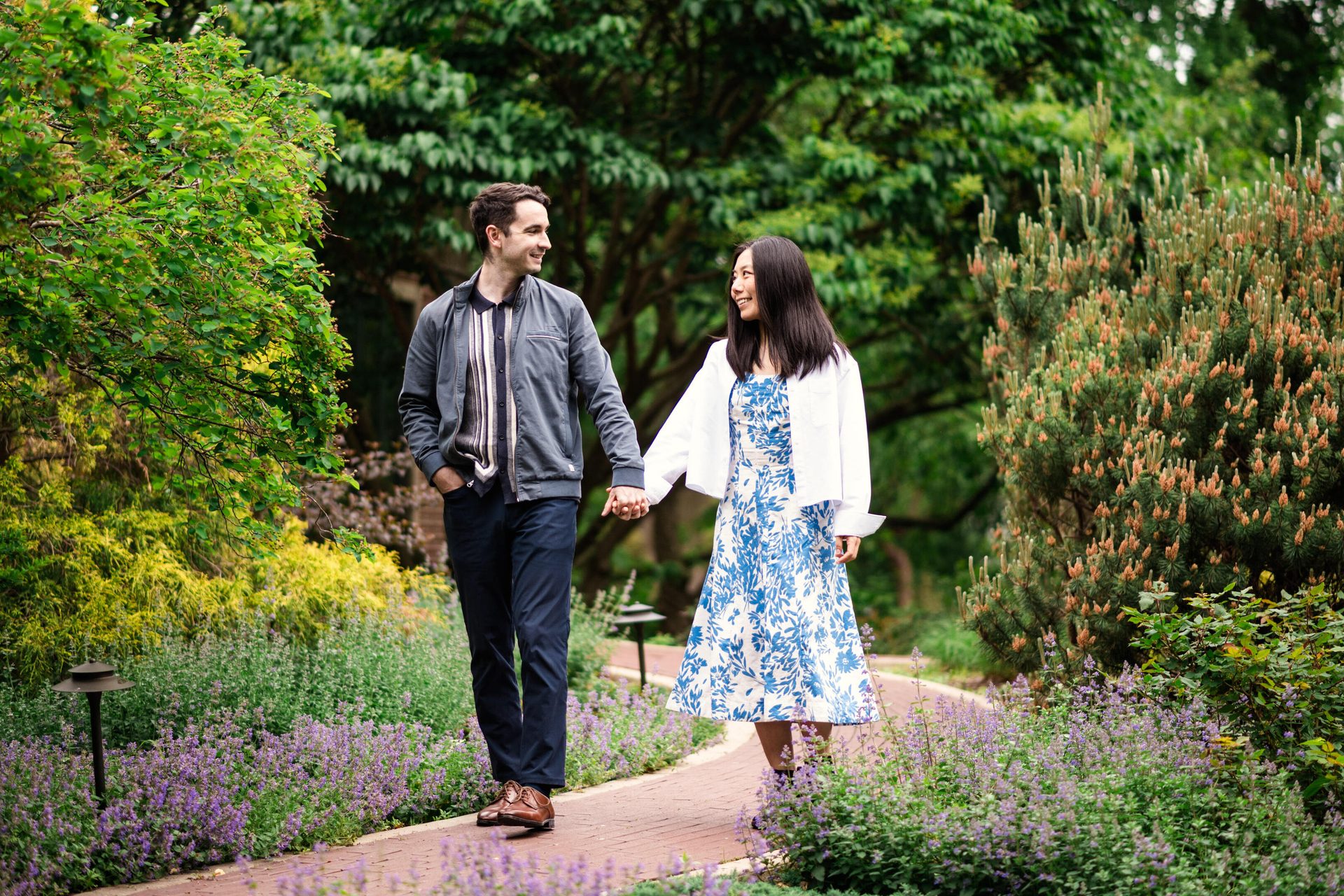 A couple walks hand-in-hand on a garden path, smiling at each other.