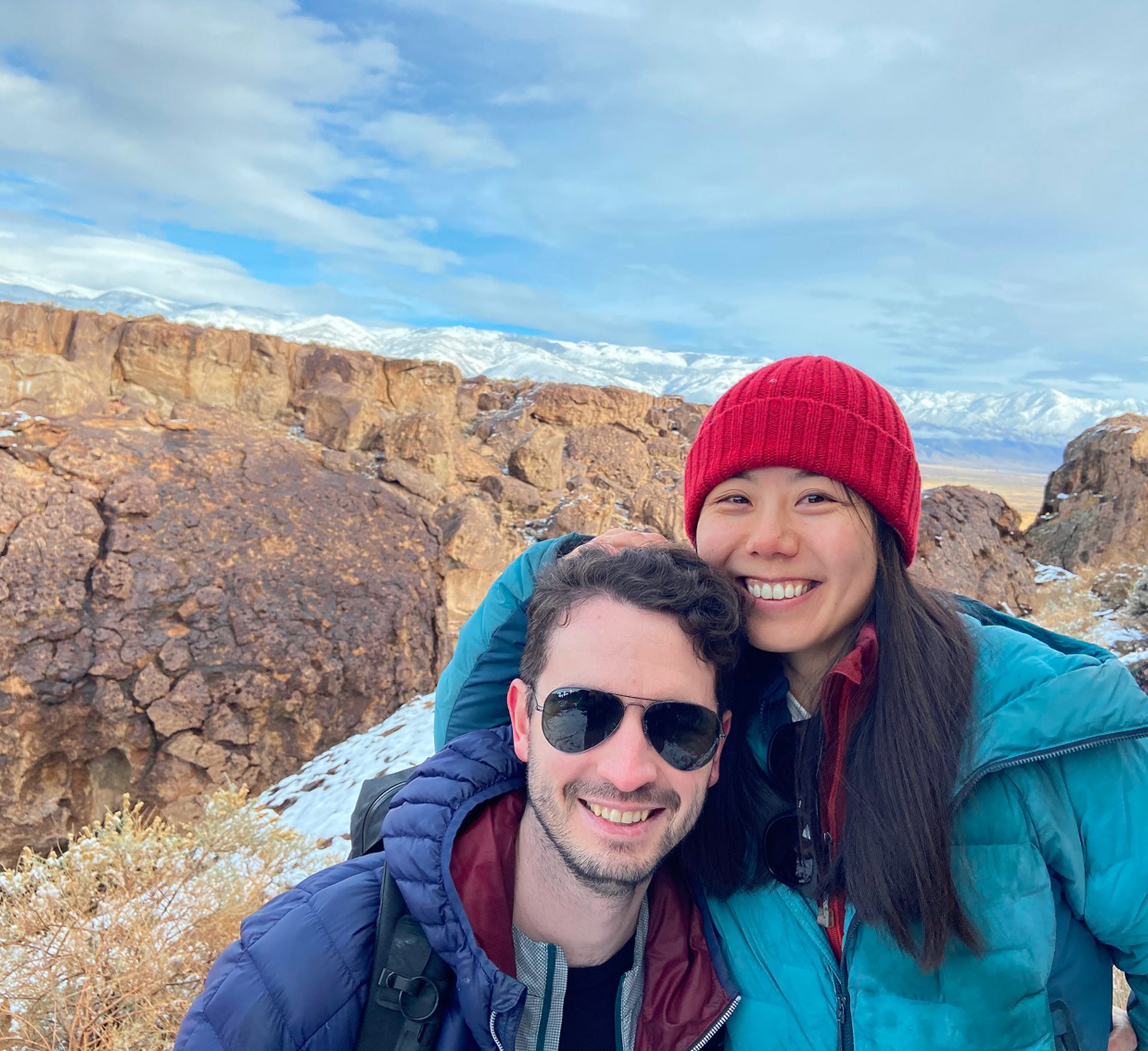 Smiling couple in winter gear against a background of snowy mountains and rocky cliffs.