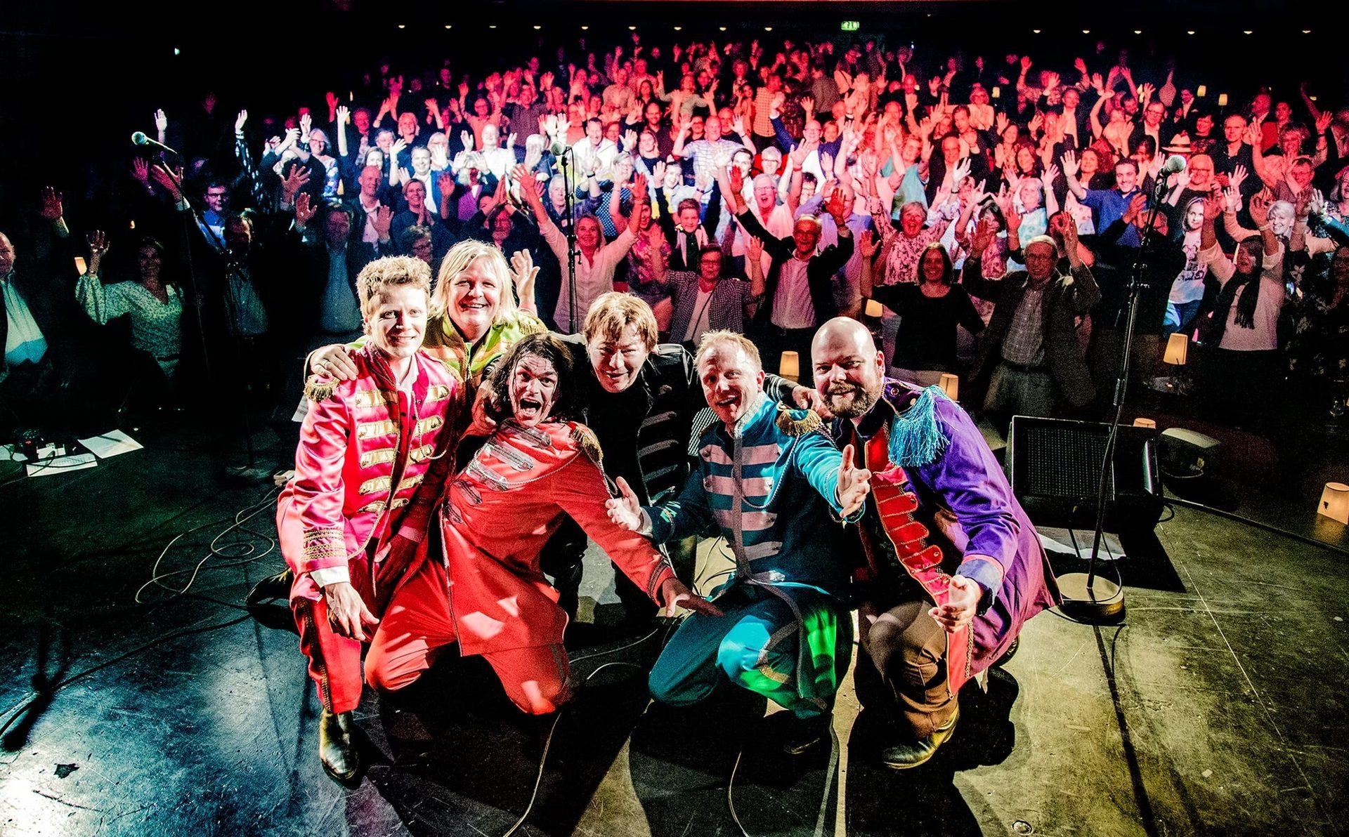 Band in colorful costumes kneeling on stage with cheering audience in background.
