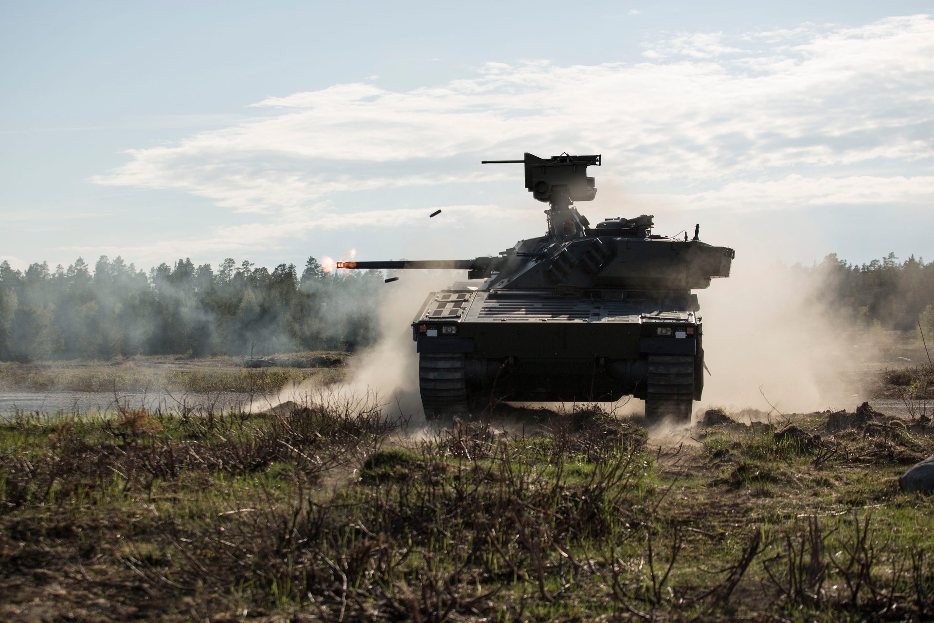 An armored vehicle fires its cannon, with muzzle flash, ejected casings, smoke, and dust in a field.