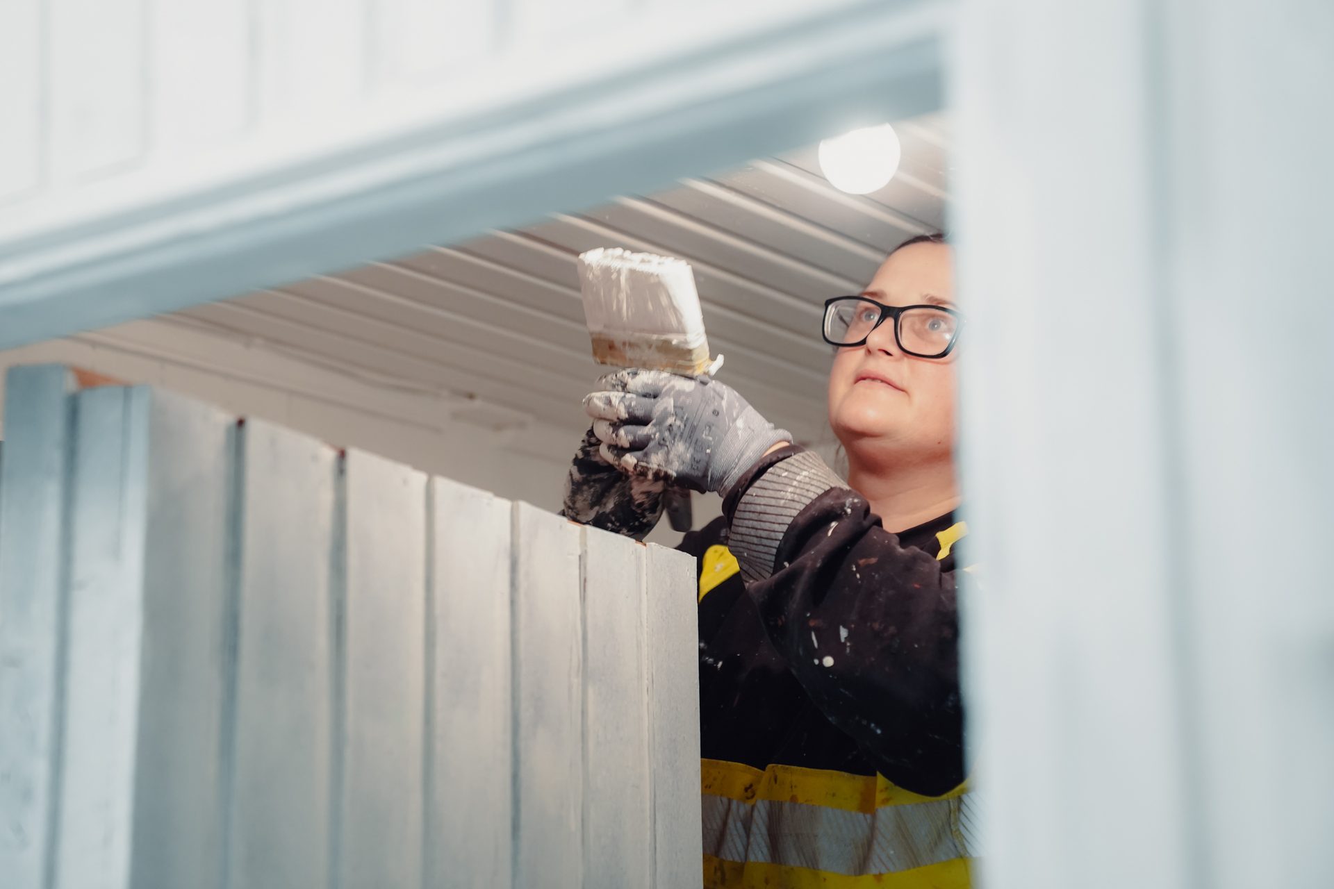 Person in glasses and gloves painting a white fence with a brush, seen from a low angle.
