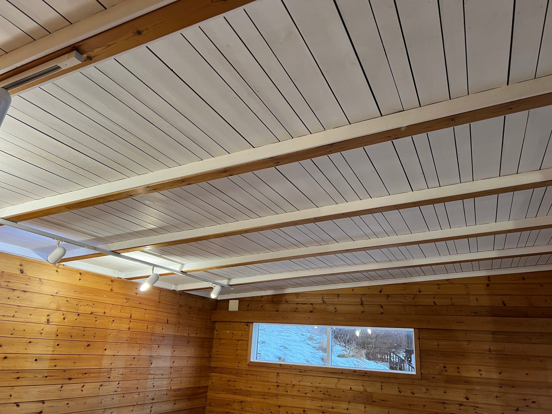 Interior shot of a room with natural wood walls, white planked ceiling, exposed beams, and a window.