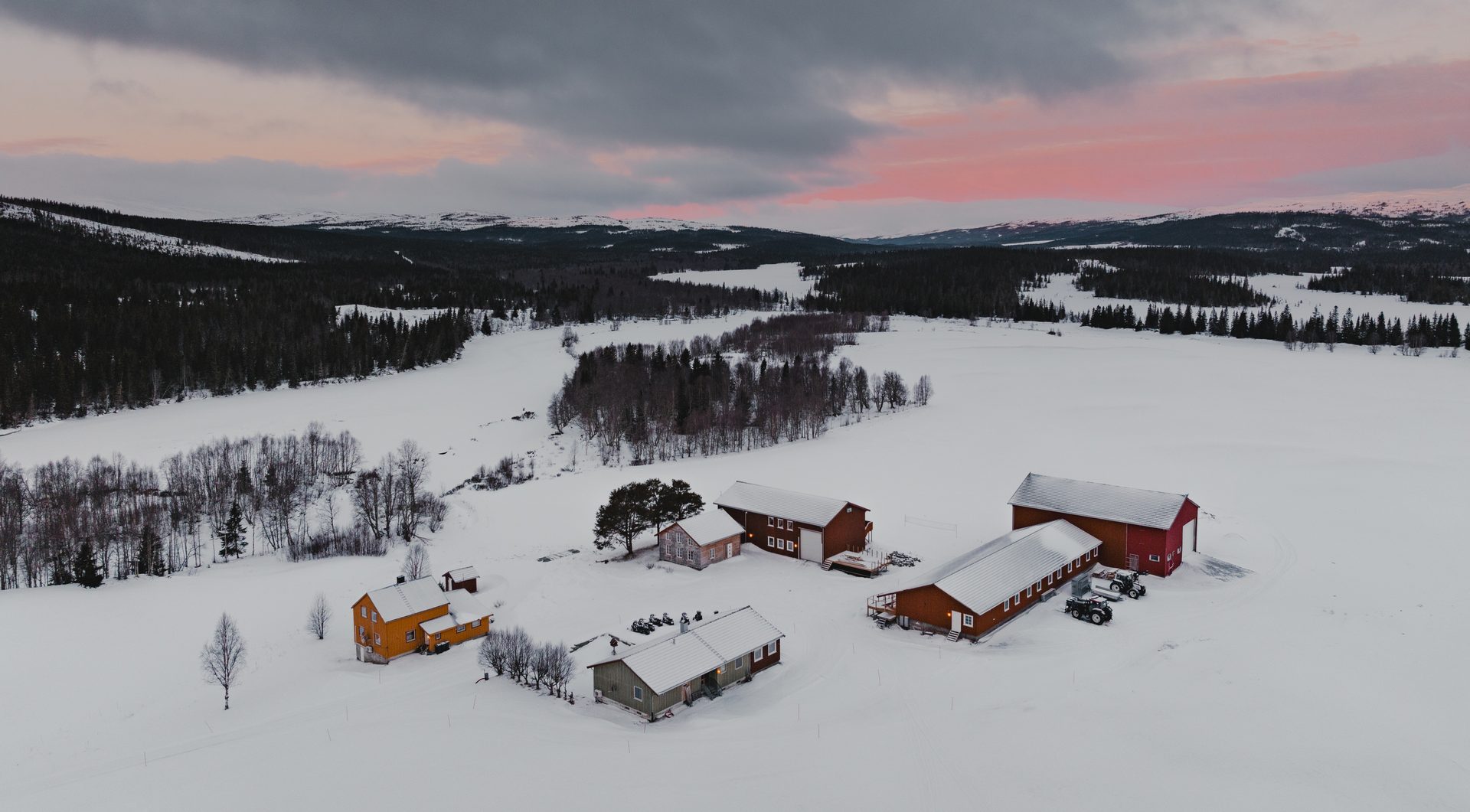 Aerial view: snowy village with colorful cabins, forests, and mountains under a pink sunset sky.
