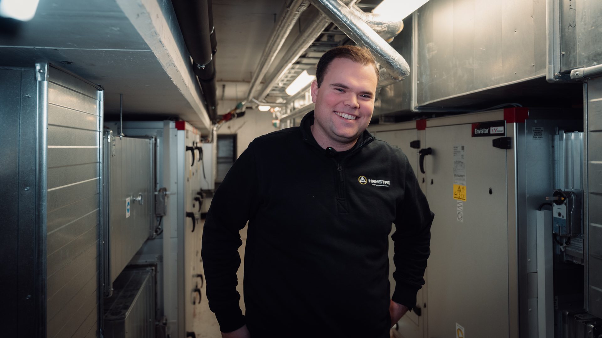 Smiling man in a black "HAMSTAD" shirt in a mechanical room with HVAC equipment and ducts.