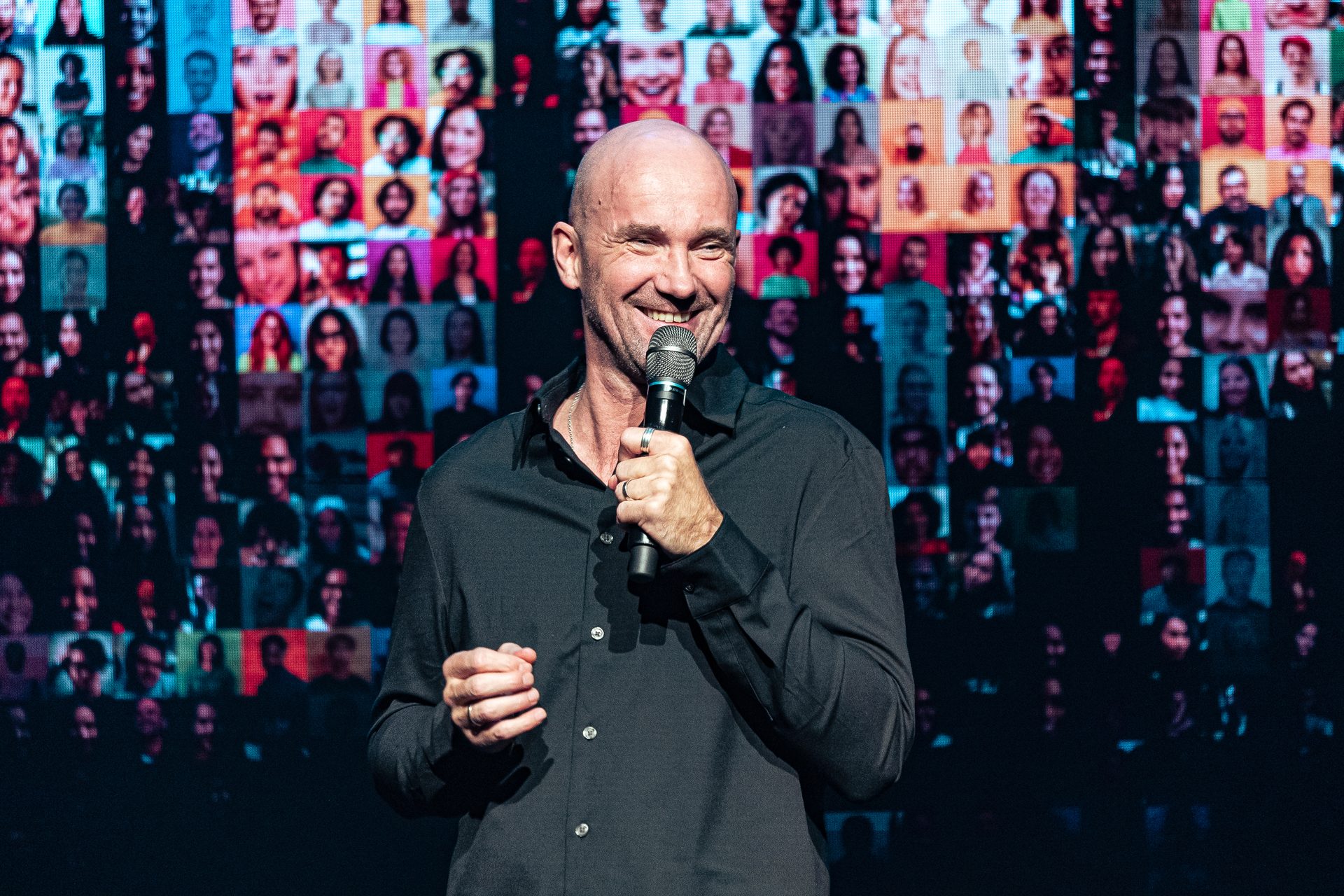 Bald man smiling, holding microphone, speaking to a virtual audience displayed on a large screen.