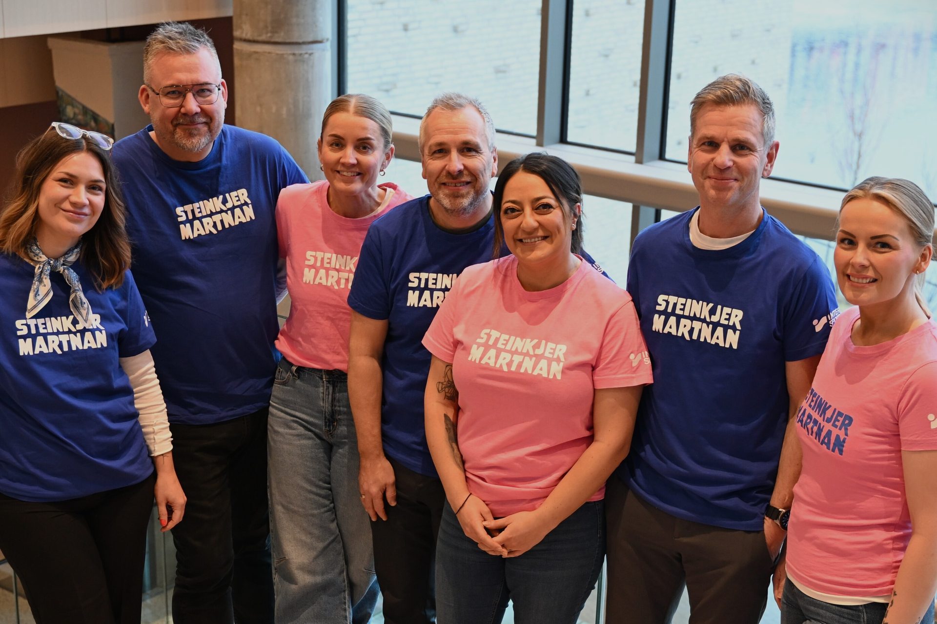Six smiling people, three men and three women, wearing blue and pink "STEINKJER MARTNAN" T-shirts.