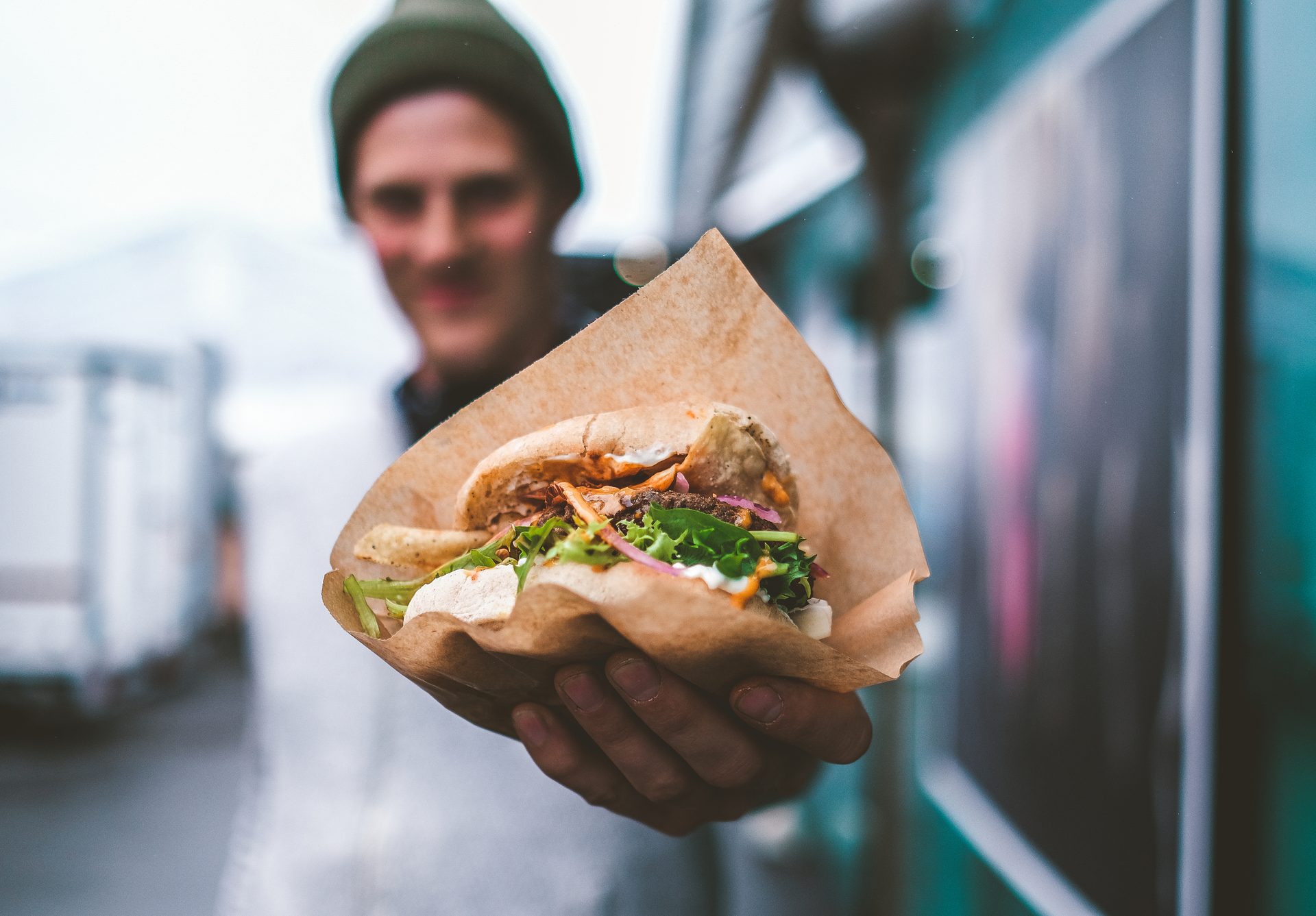 Blurred person holds out a pita sandwich, wrapped in paper.