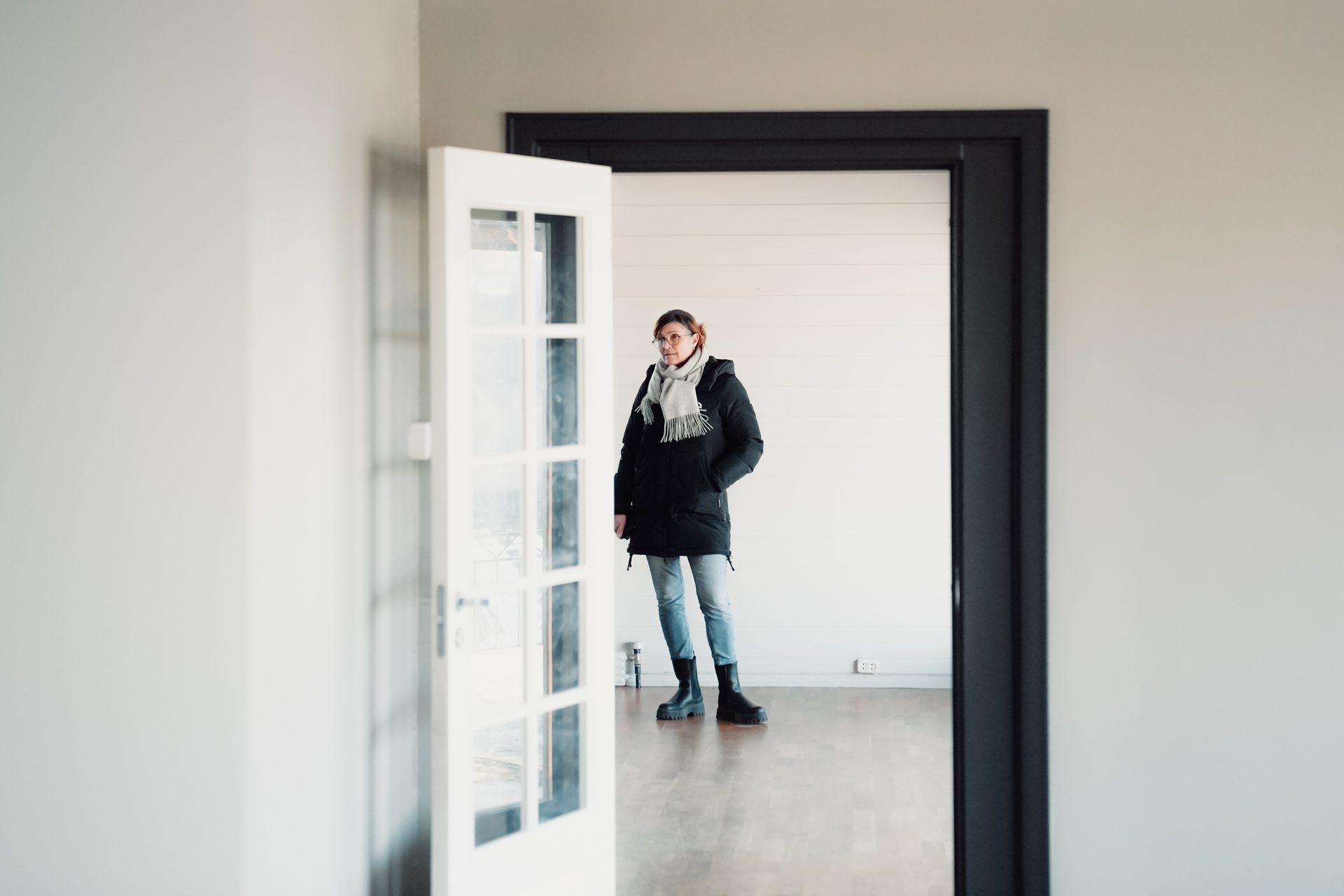 Woman in a black winter coat and grey scarf standing in an empty room, looking around.