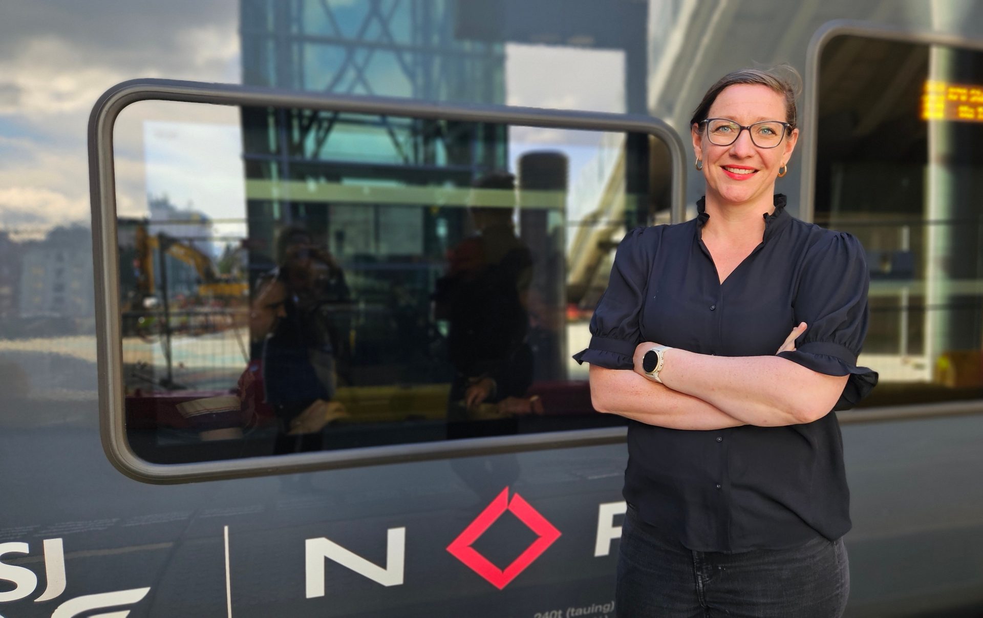 Smiling woman in glasses and dark shirt, arms crossed, in front of a train with NOR logo.