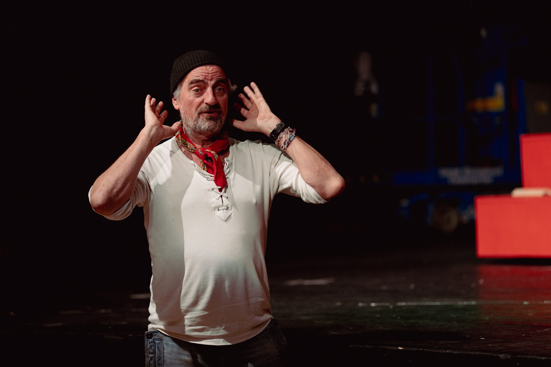 Bearded man in beanie and red scarf, hands to ears, expressive on a dark stage.