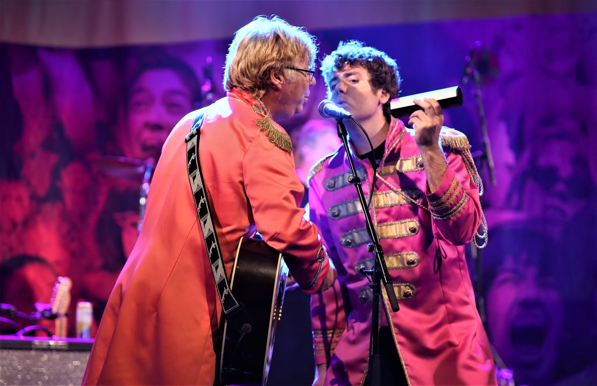 Two musicians in pink and orange ornate jackets perform on a stage with a guitar and harmonica.