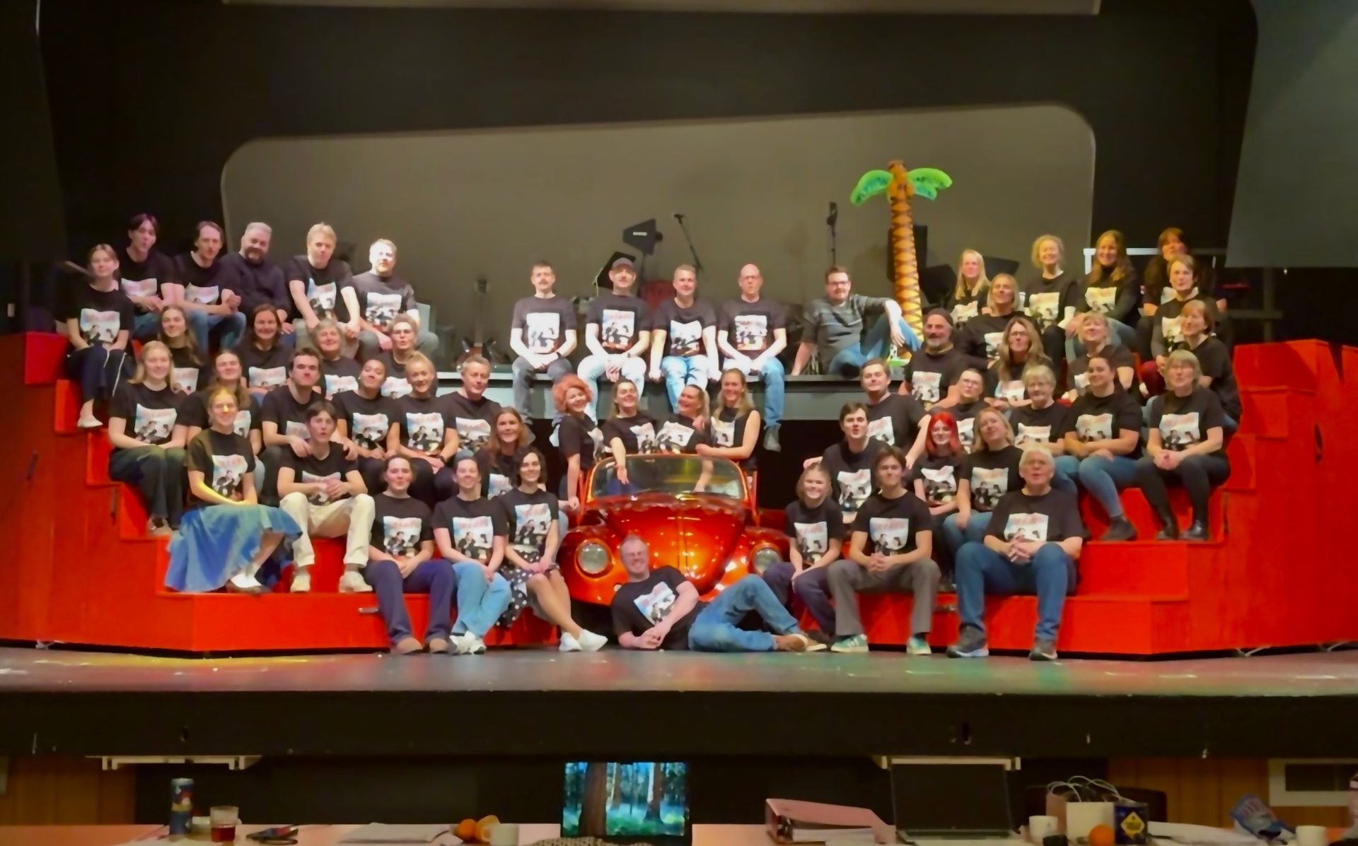 Large group of people in black shirts posing on a stage with red stairs and a red car.