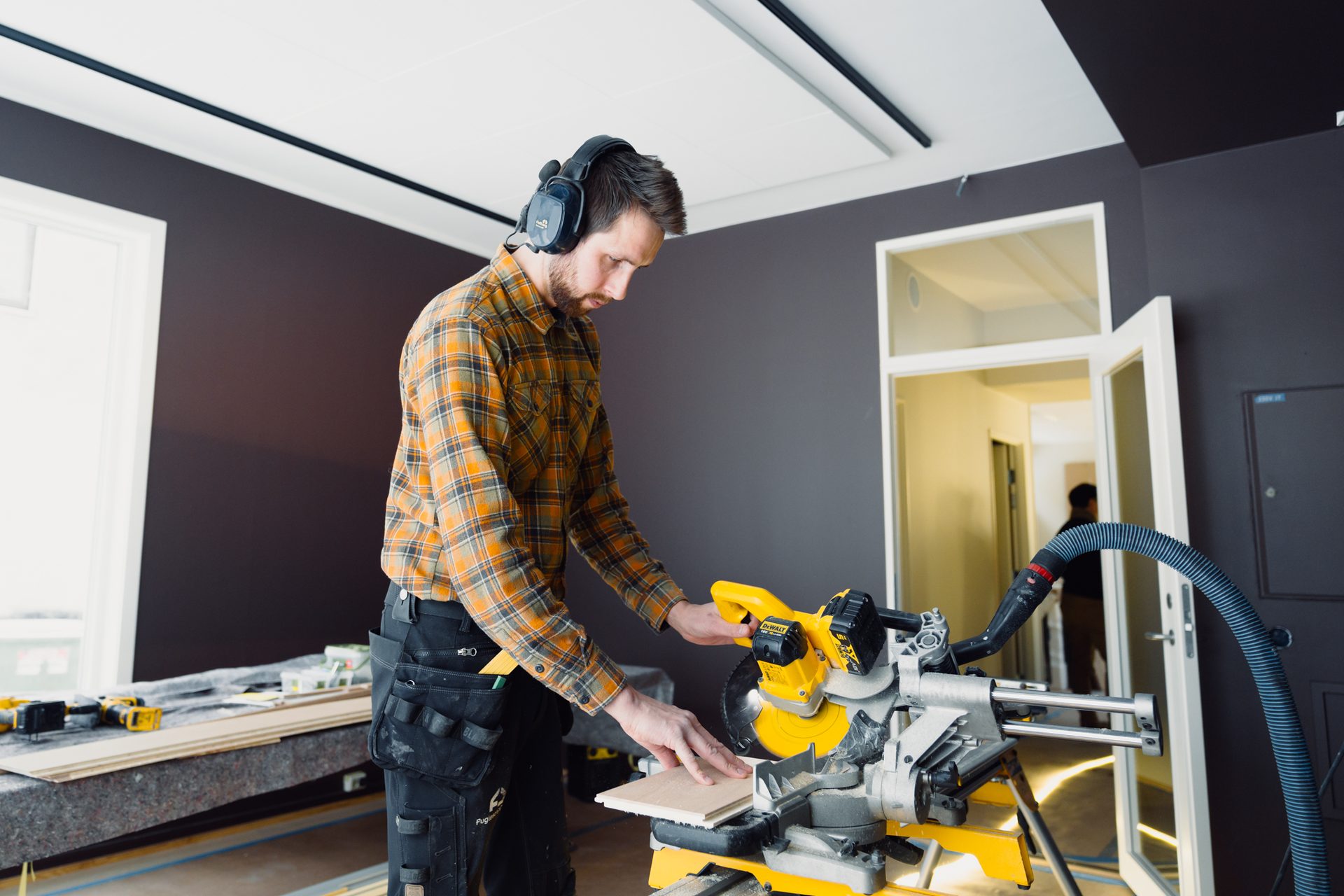 Man in plaid shirt and safety headphones cutting wood with a miter saw in a room undergoing renovation.