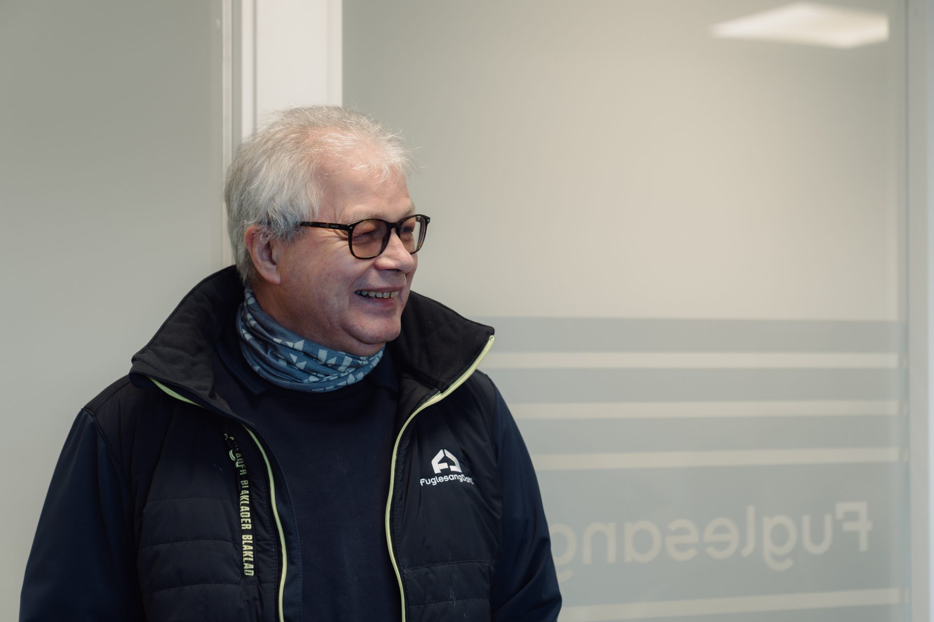 Older man with gray hair and glasses smiling, wearing a black work jacket and patterned neck gaiter.