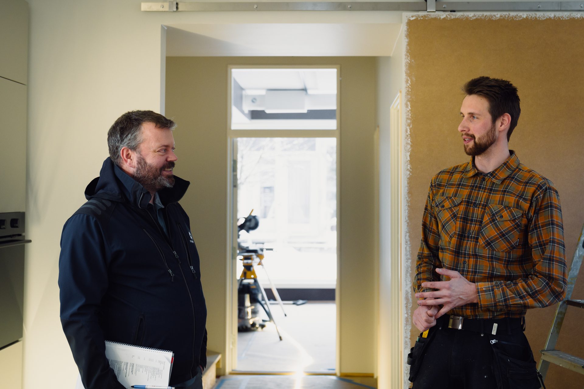 Two men, possibly contractors, discussing in a partially renovated indoor space with a doorway in the background.