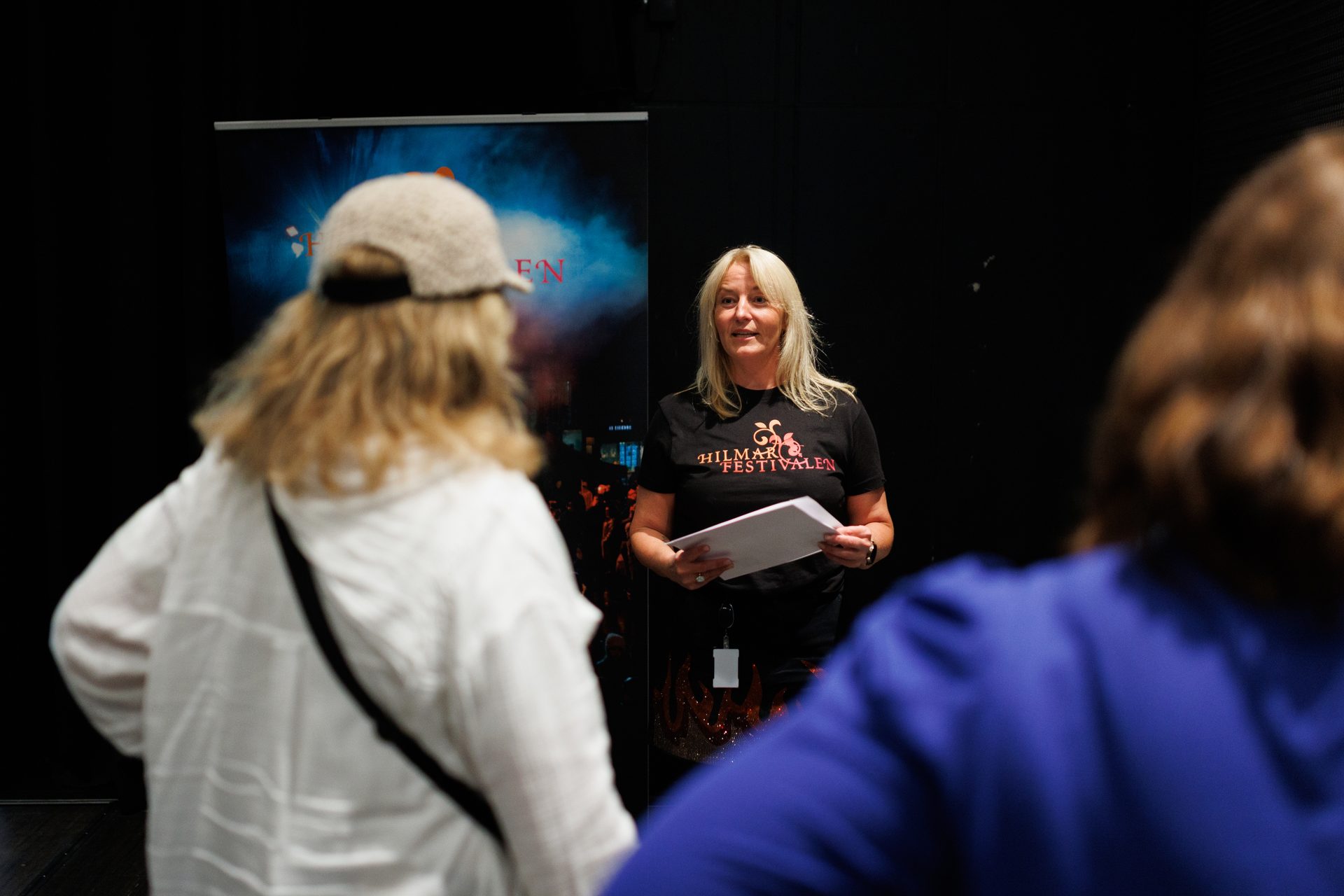 A woman in a black "Hilmar Festivalen" shirt holds papers, speaking to a blurred audience.