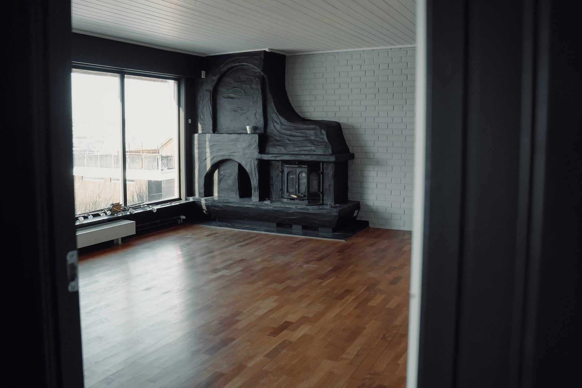 An empty room featuring a large, dark, sculptural fireplace, a bright window, and a polished wooden floor.