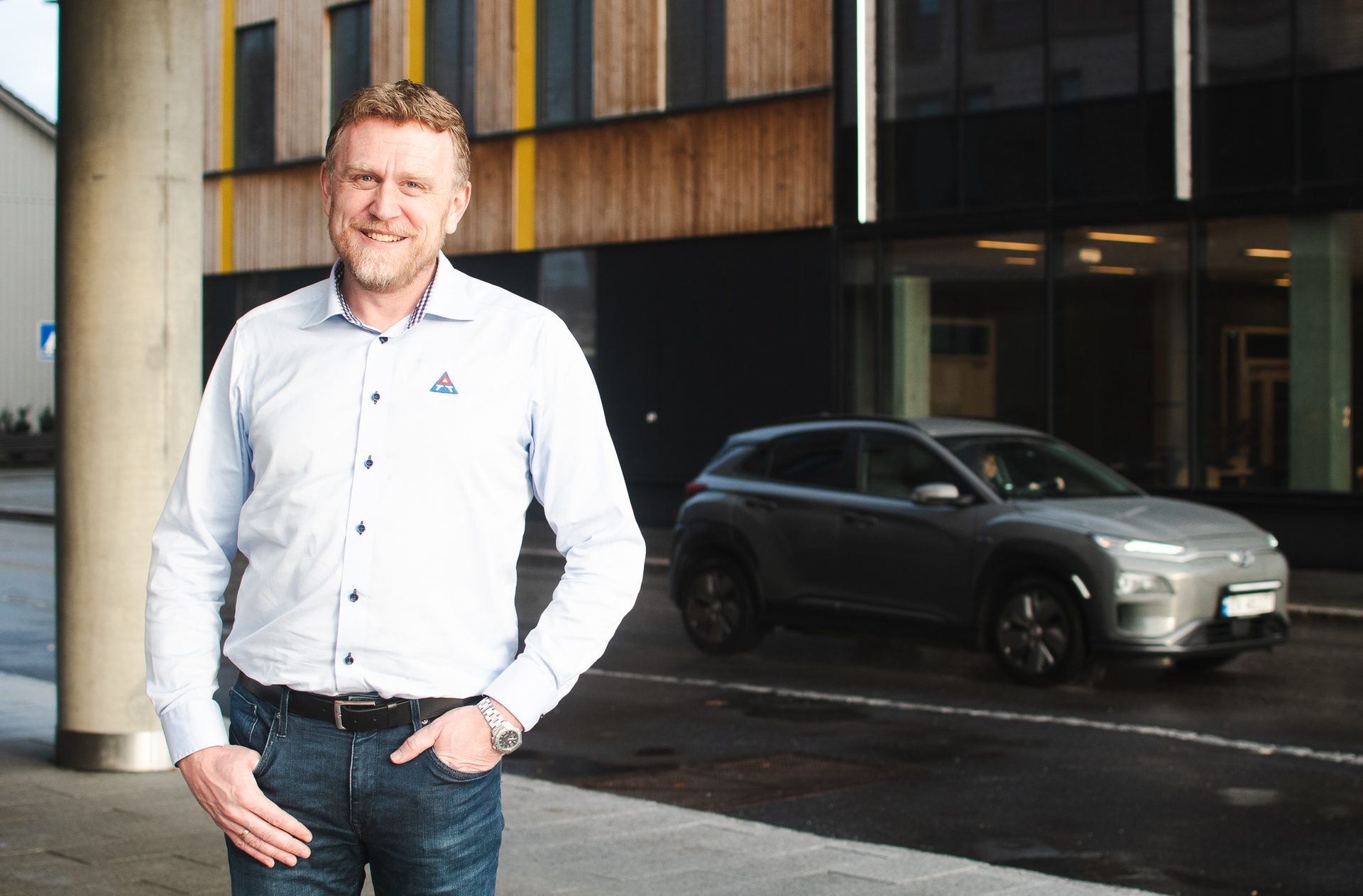 A smiling man in a light blue shirt and jeans stands on a sidewalk, with a silver electric car behind him.