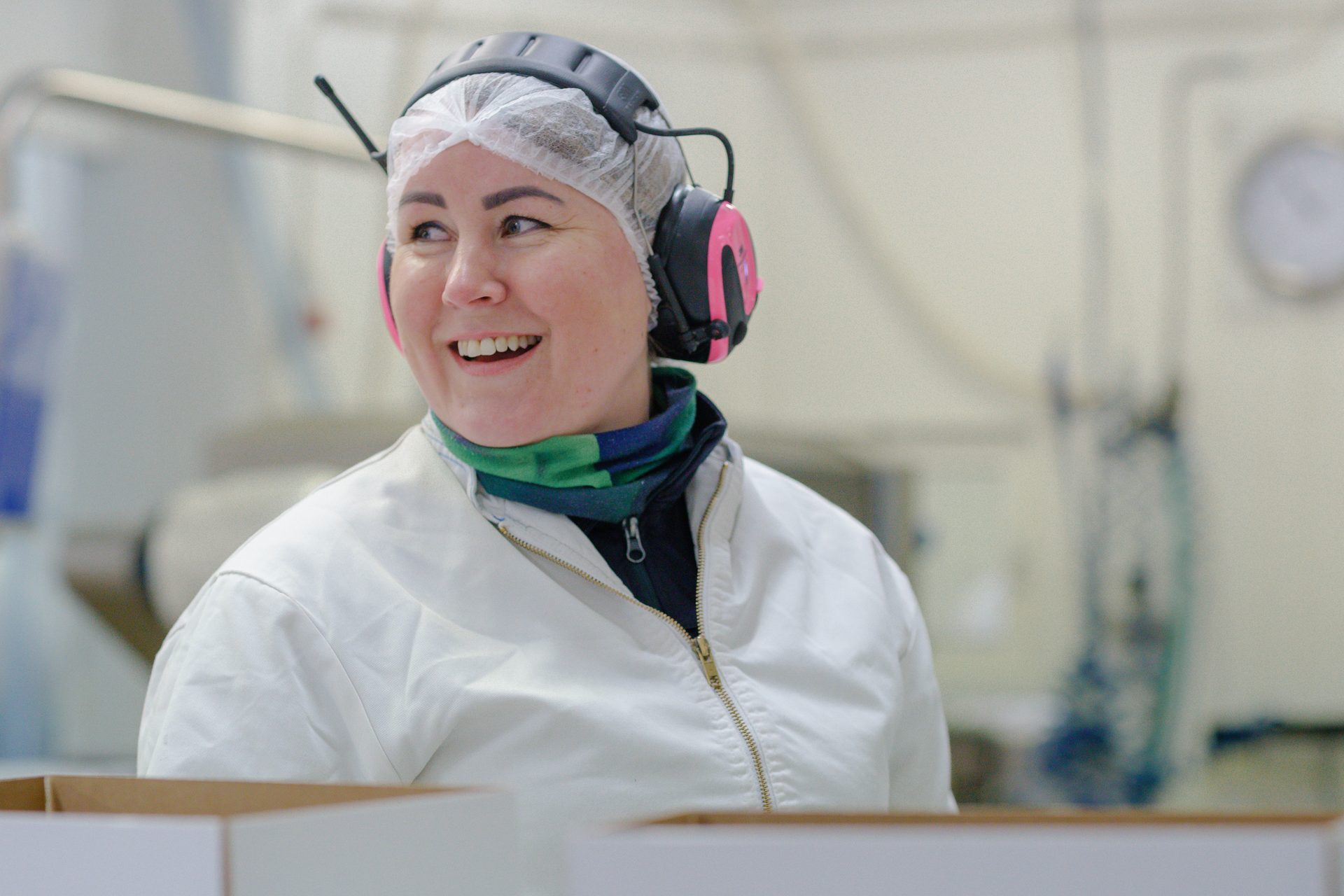 Smiling woman in a hairnet, pink ear protection, and white protective coat in a factory.