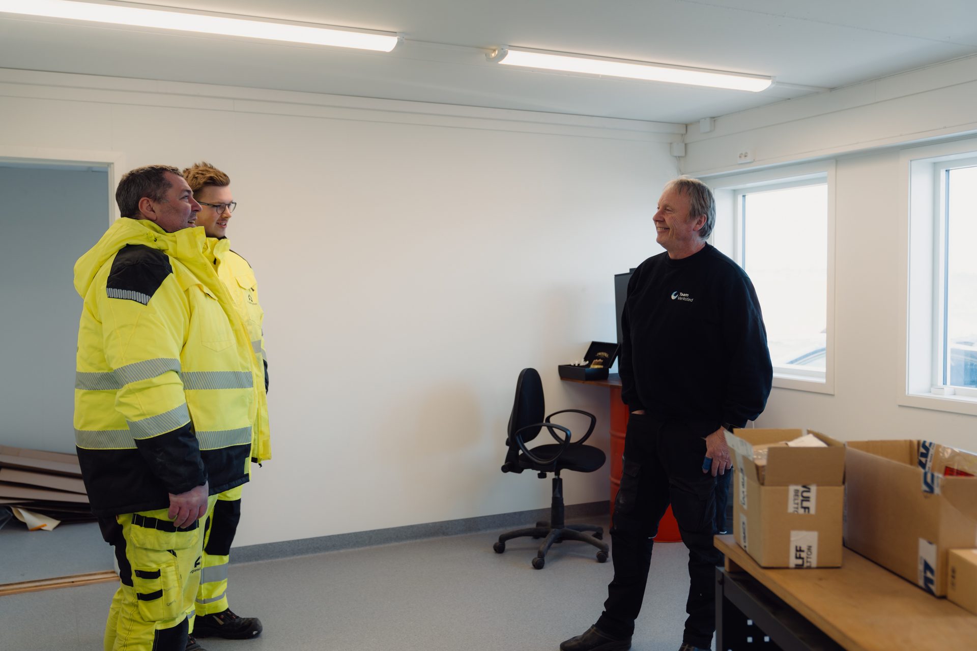 Three men, two in high-visibility gear and one in black, conversing in a bright, modern room.
