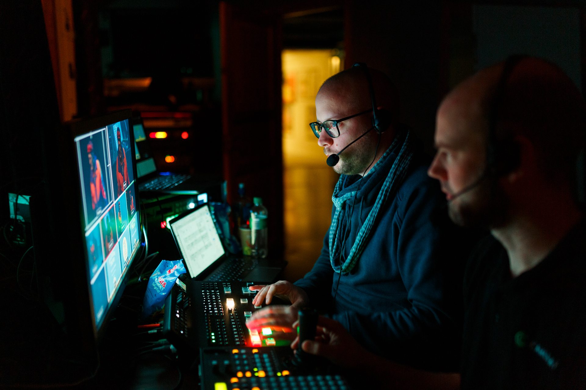 Two men in headsets control multi-screen video production in a dark room.