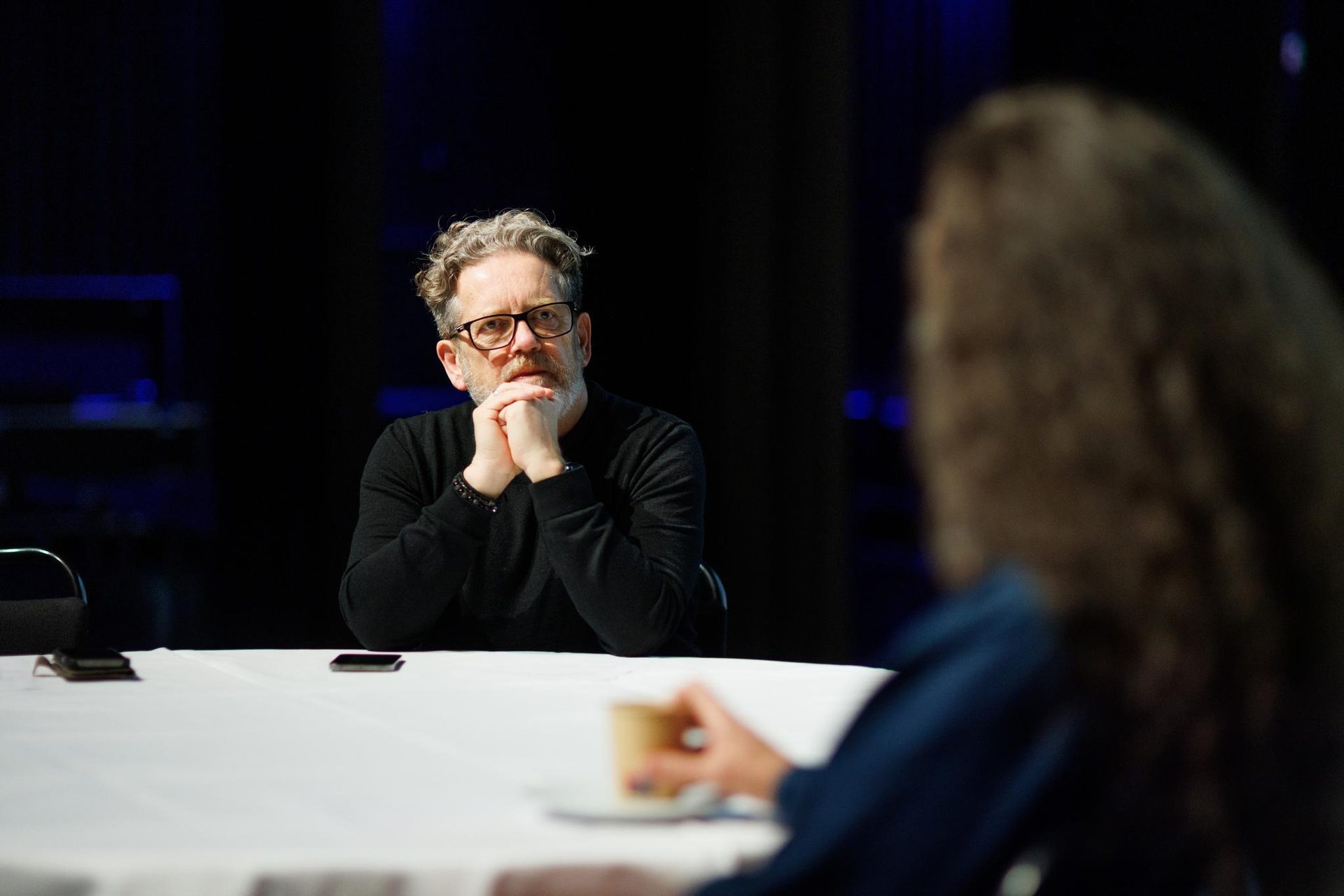 Man with glasses, beard, hands clasped, looking at person across table with cup.