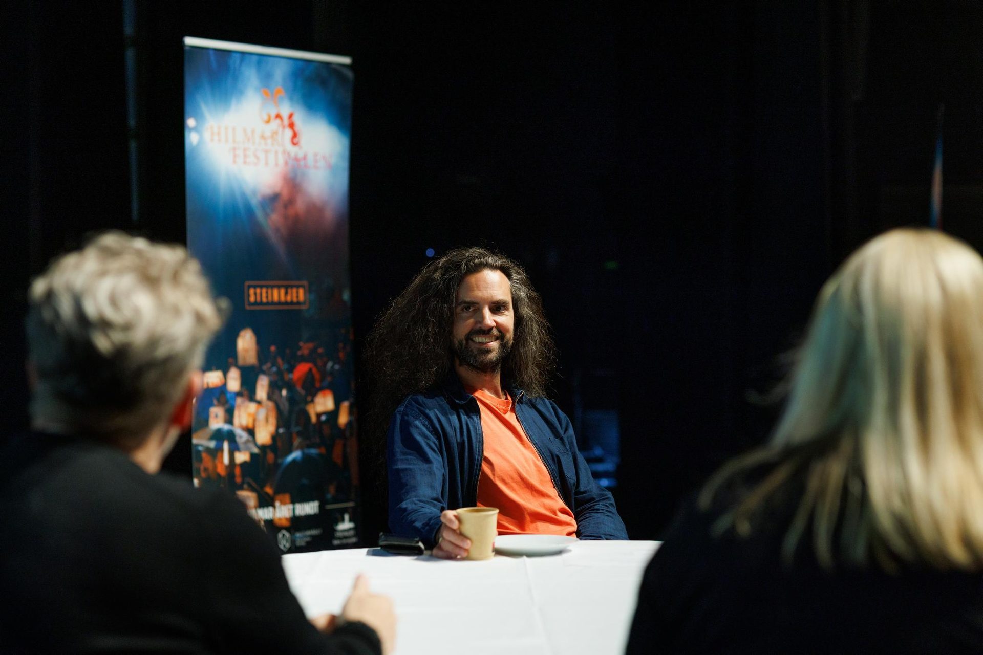 Smiling man with long hair and beard at a table between two people, 'Hilmar Festivalen' banner behind.