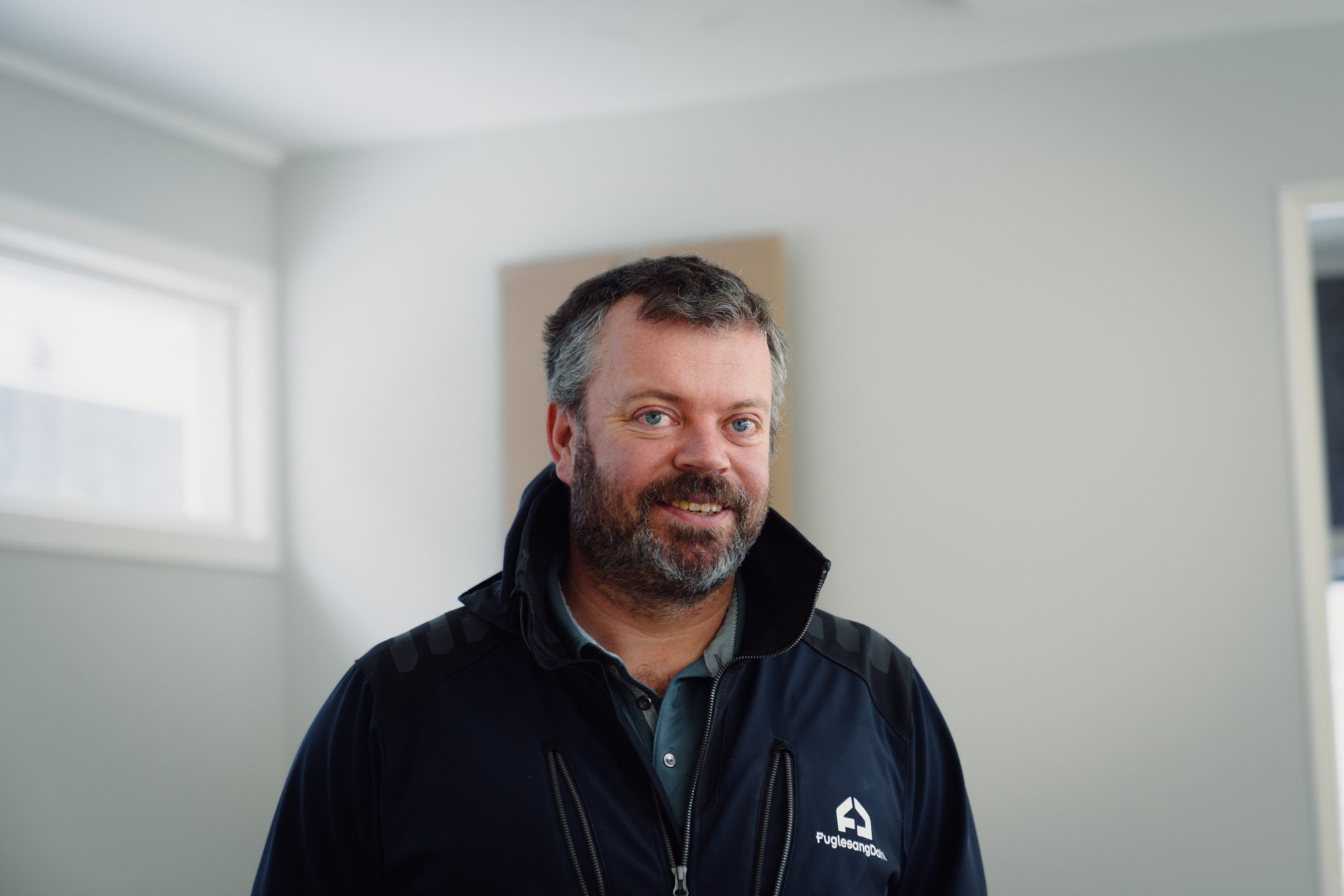A man with a beard and grey hair, wearing a dark jacket with a logo, smiling at the camera.