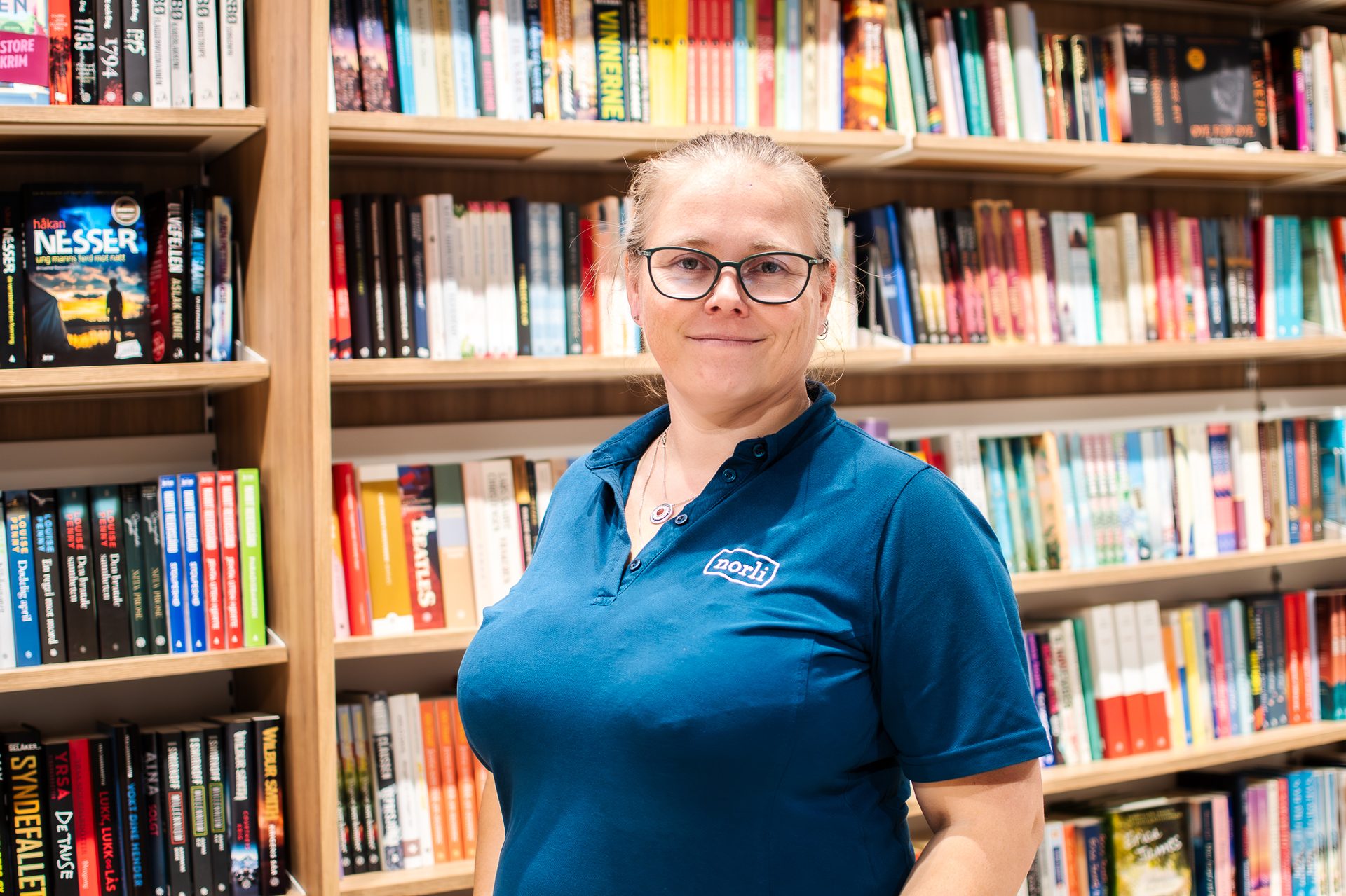 Smiling woman in blue Norli polo shirt and glasses in a bookstore.