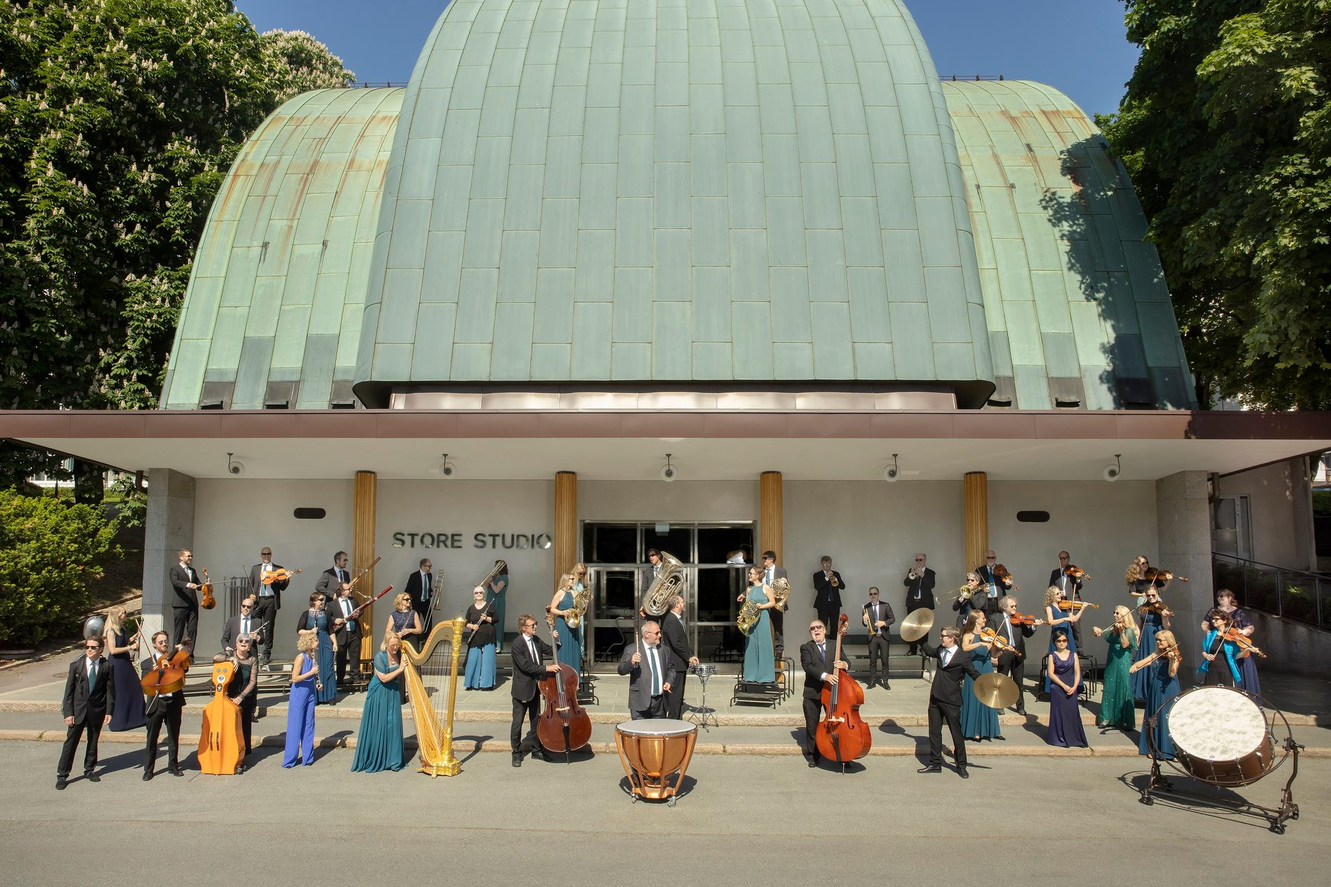 A large orchestra with various instruments stands outside a building with a curved green roof.