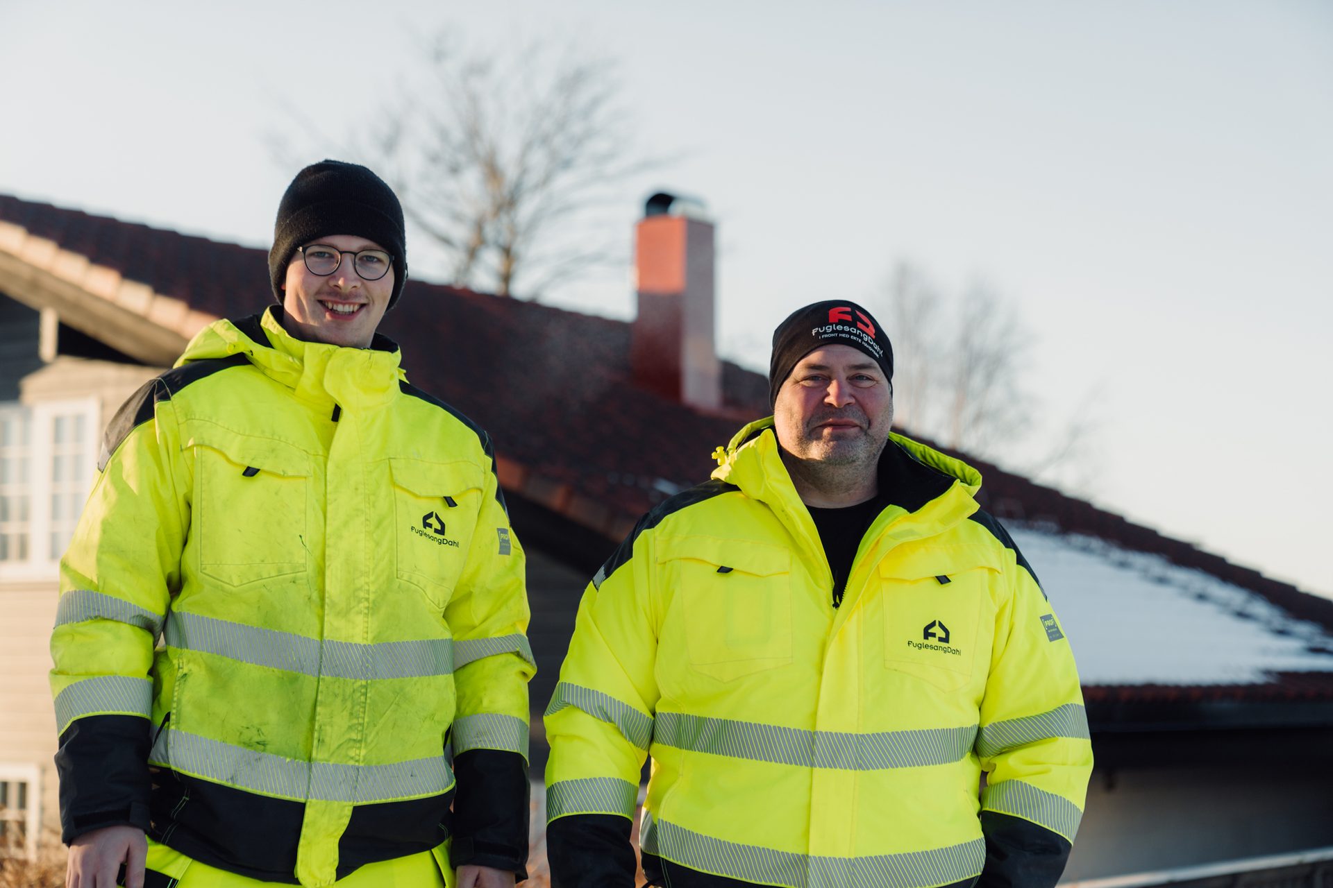 Two men in high-visibility jackets and beanies smiling outdoors.