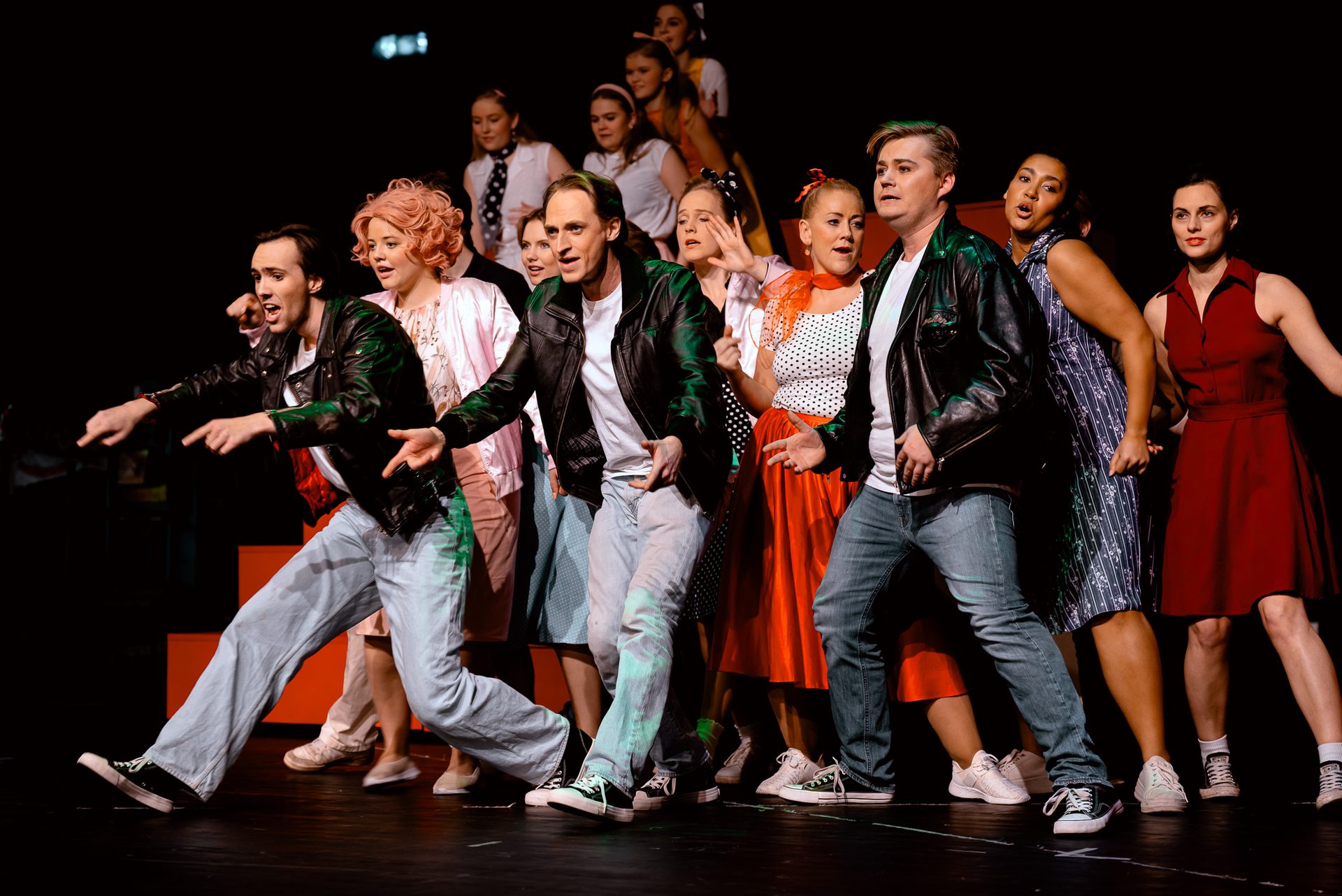 A lively group of performers in 1950s-style clothing on a dark stage, actively posing and pointing.