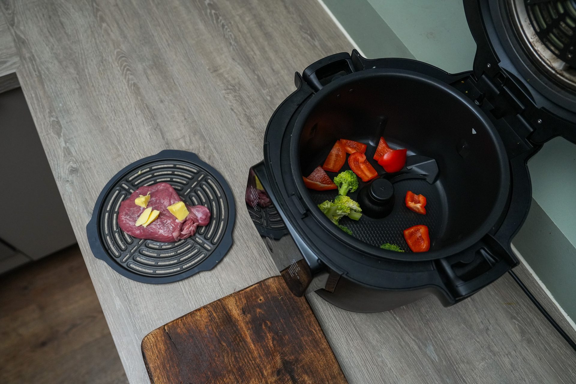 Raw meat on a grill plate and vegetables in an open air fryer next to a cutting board.