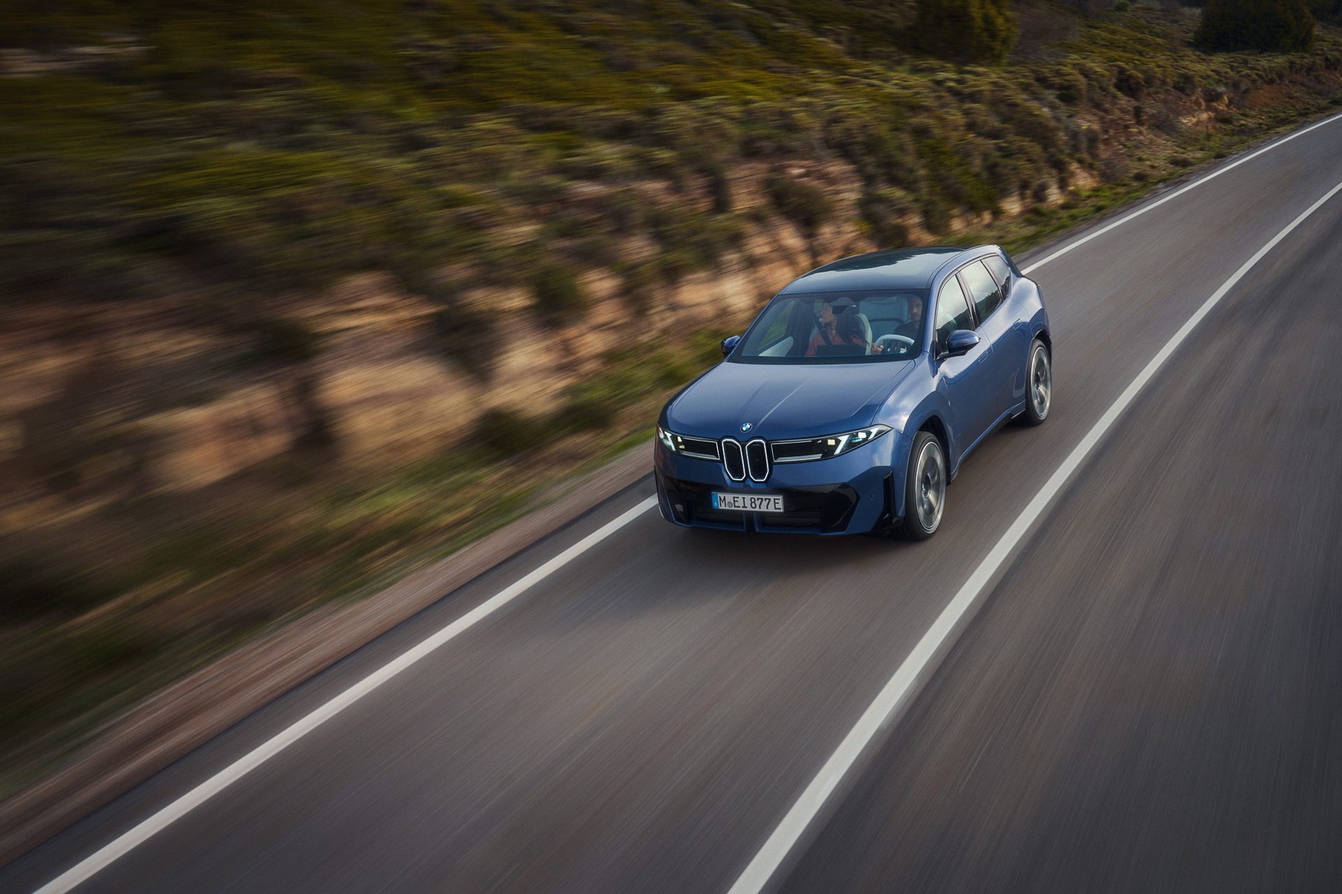 Blue car on a highway with motion blur, driving past a brushy hillside.