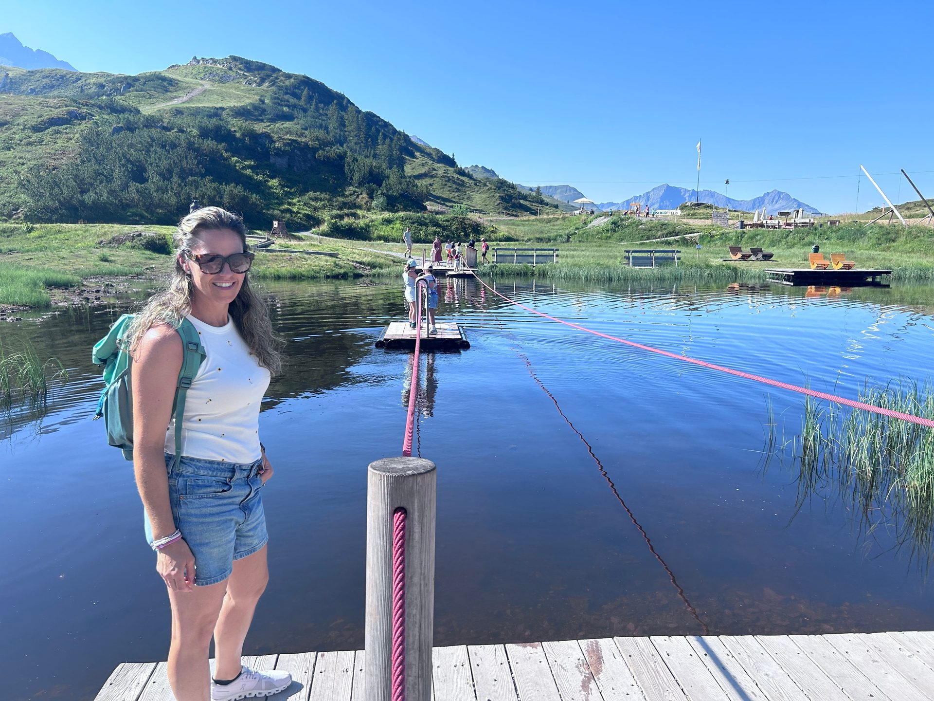 Woman standing on a wooden pier next to a mountain lake with people on floating platforms in the background.