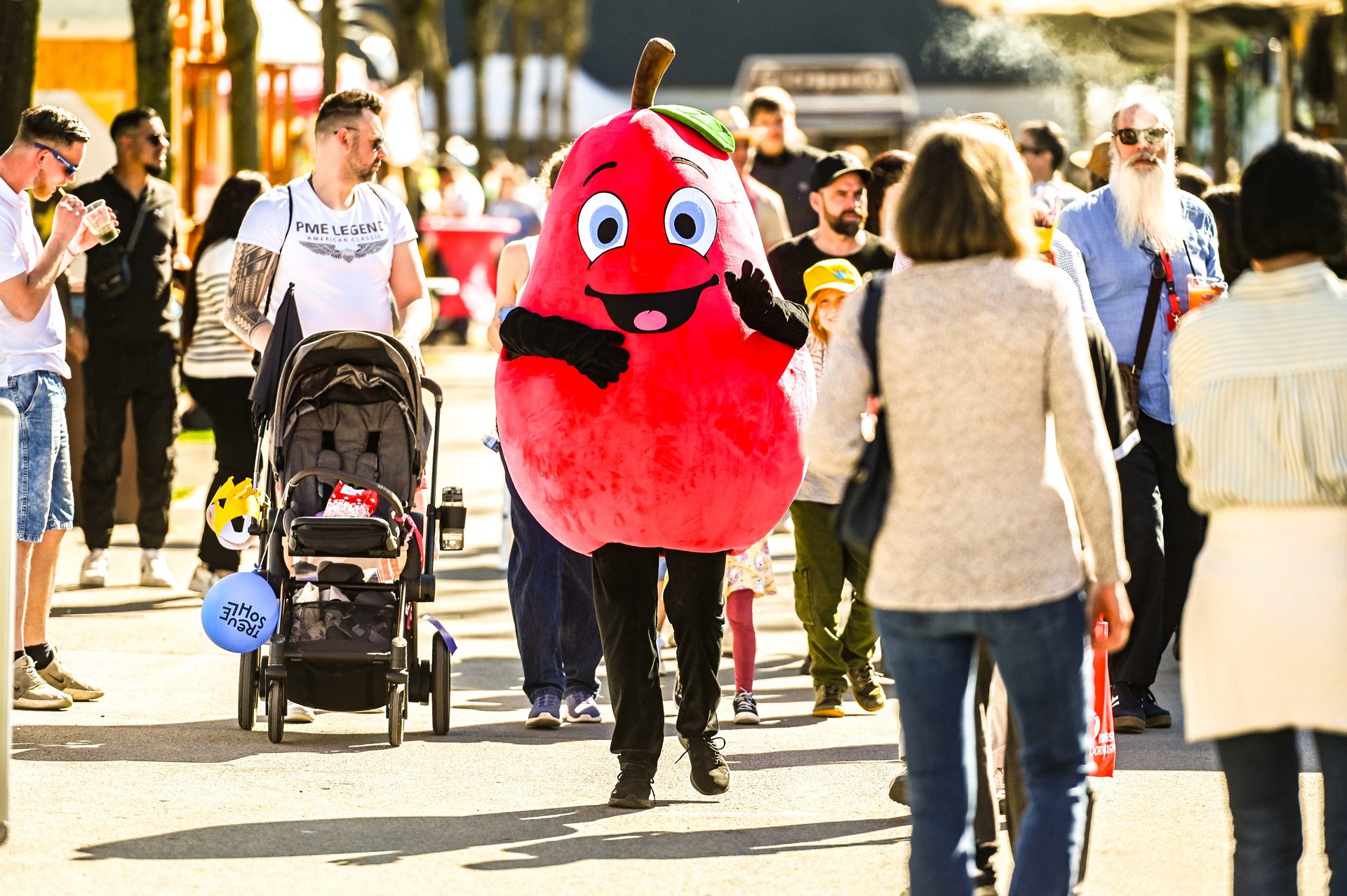 A person in a cheerful red pear mascot costume waves in a sunny outdoor crowd with a man pushing a stroller.