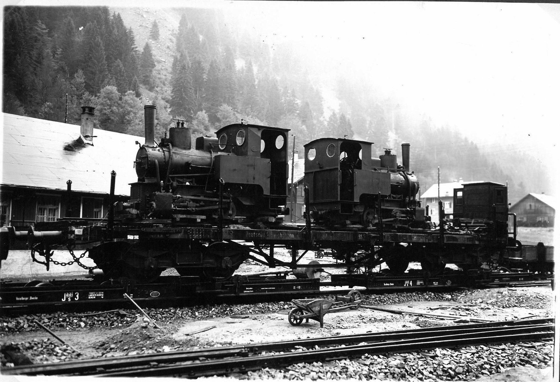 Two small steam locomotives on flatbed railcars in a mountain setting with buildings and forests.