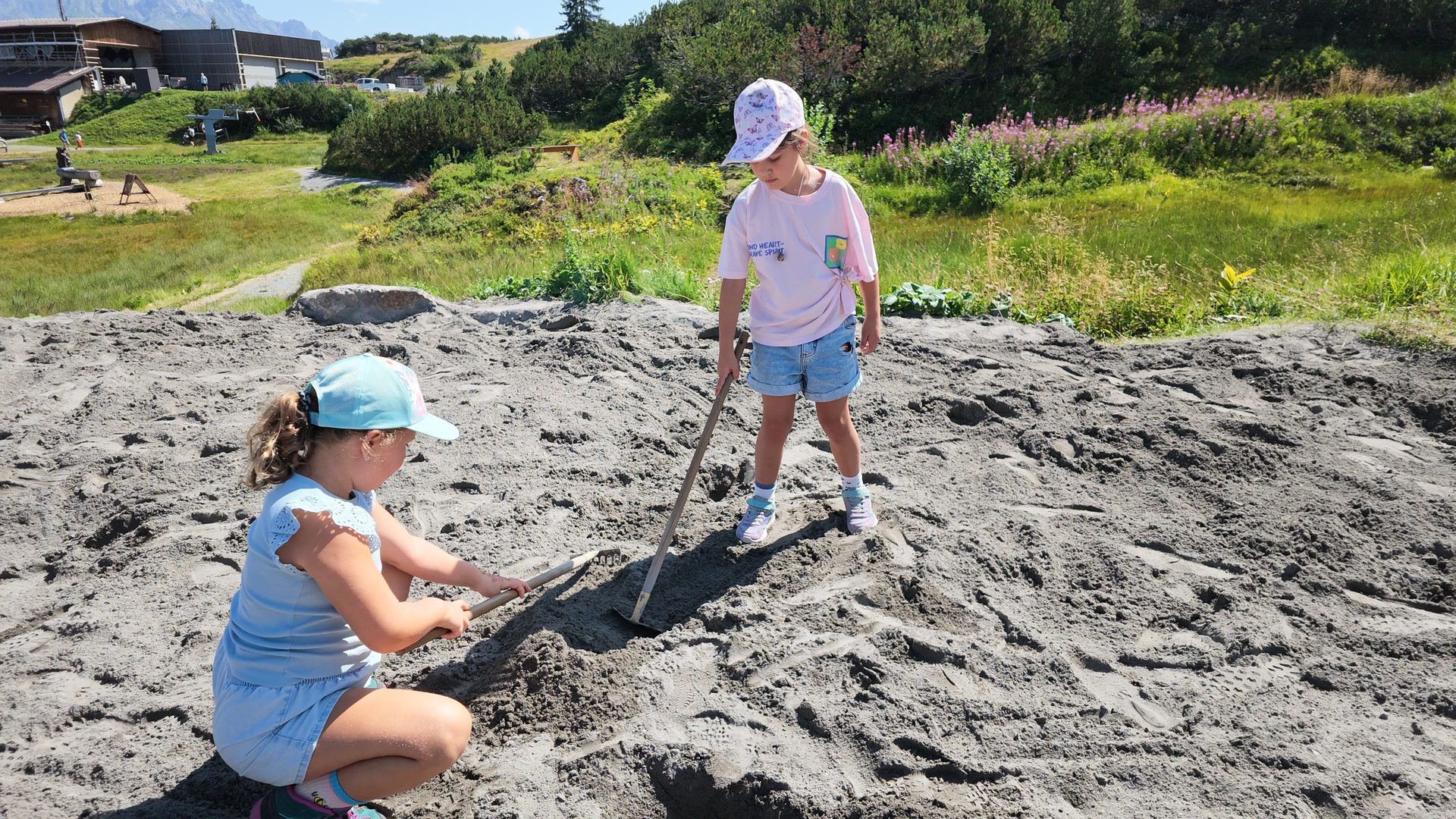 Two young girls playing in a large sandpit with rakes on a sunny day, with green hills behind them.