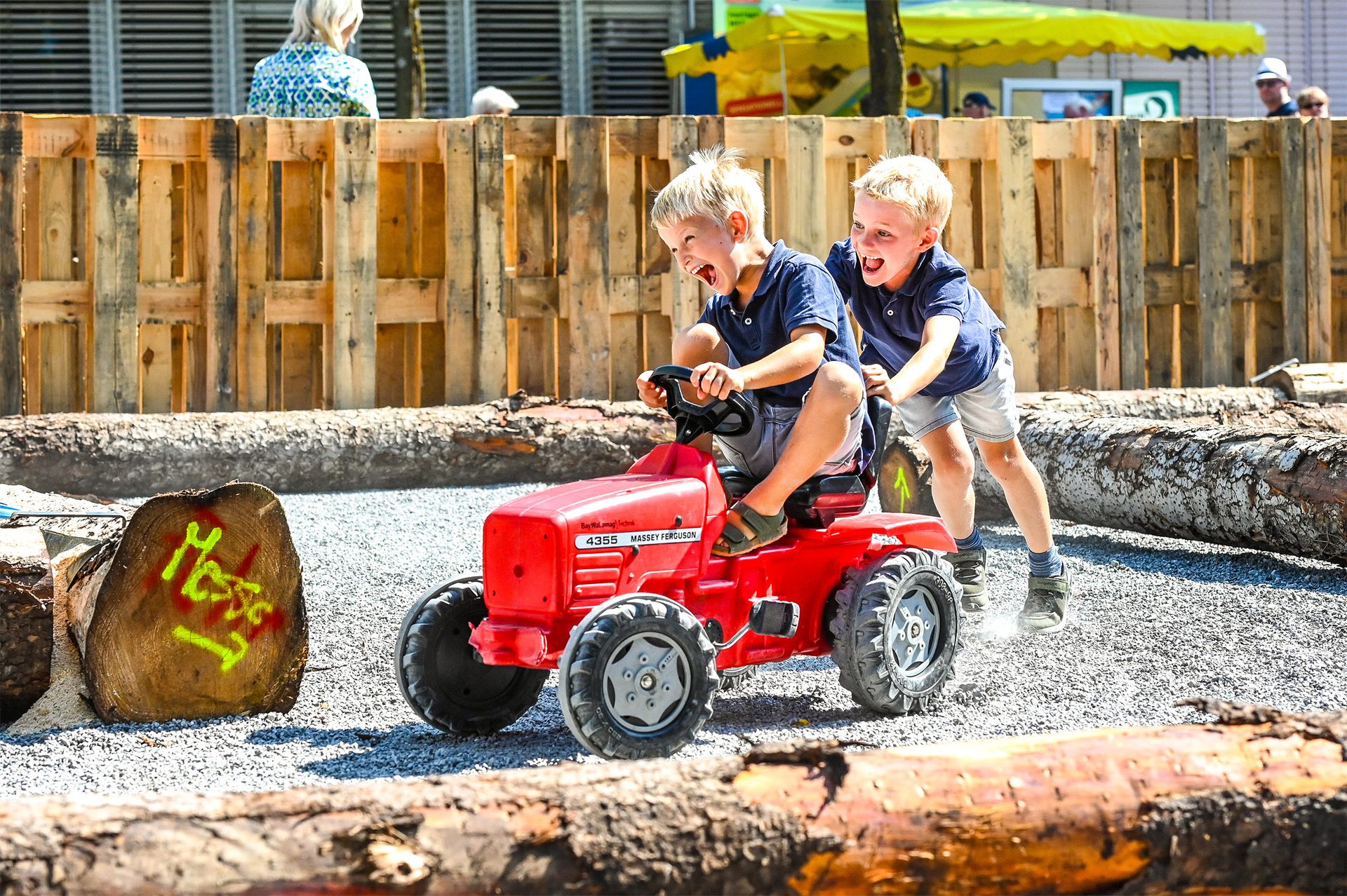 Two excited boys playing with a red toy tractor outdoors, one driving and the other pushing.