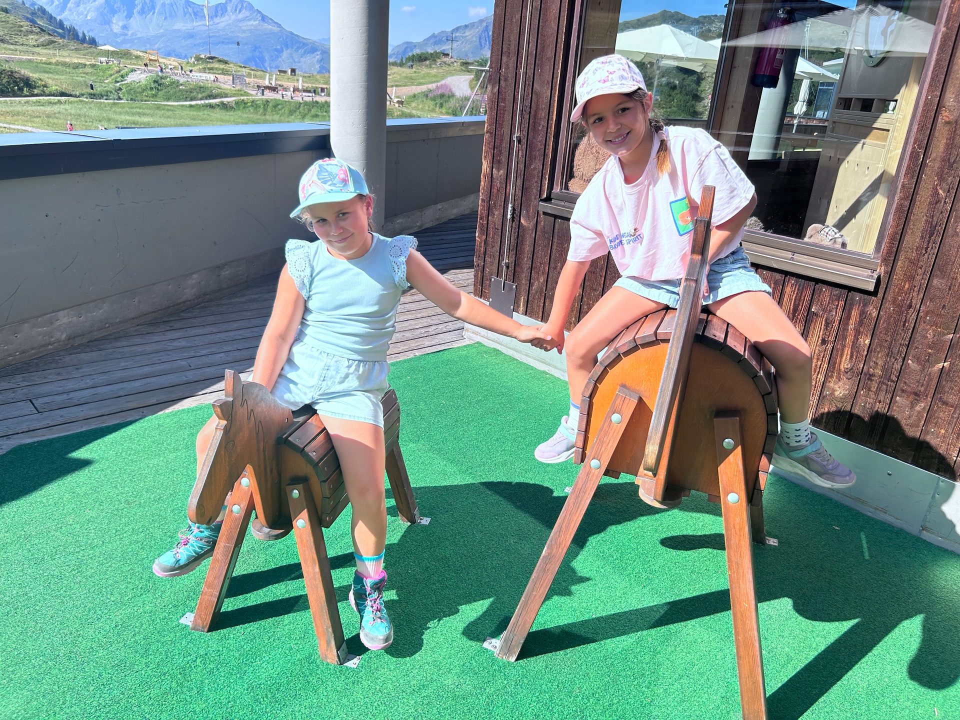 Two smiling girls riding wooden horses, holding hands, with a scenic mountain backdrop on a sunny day.