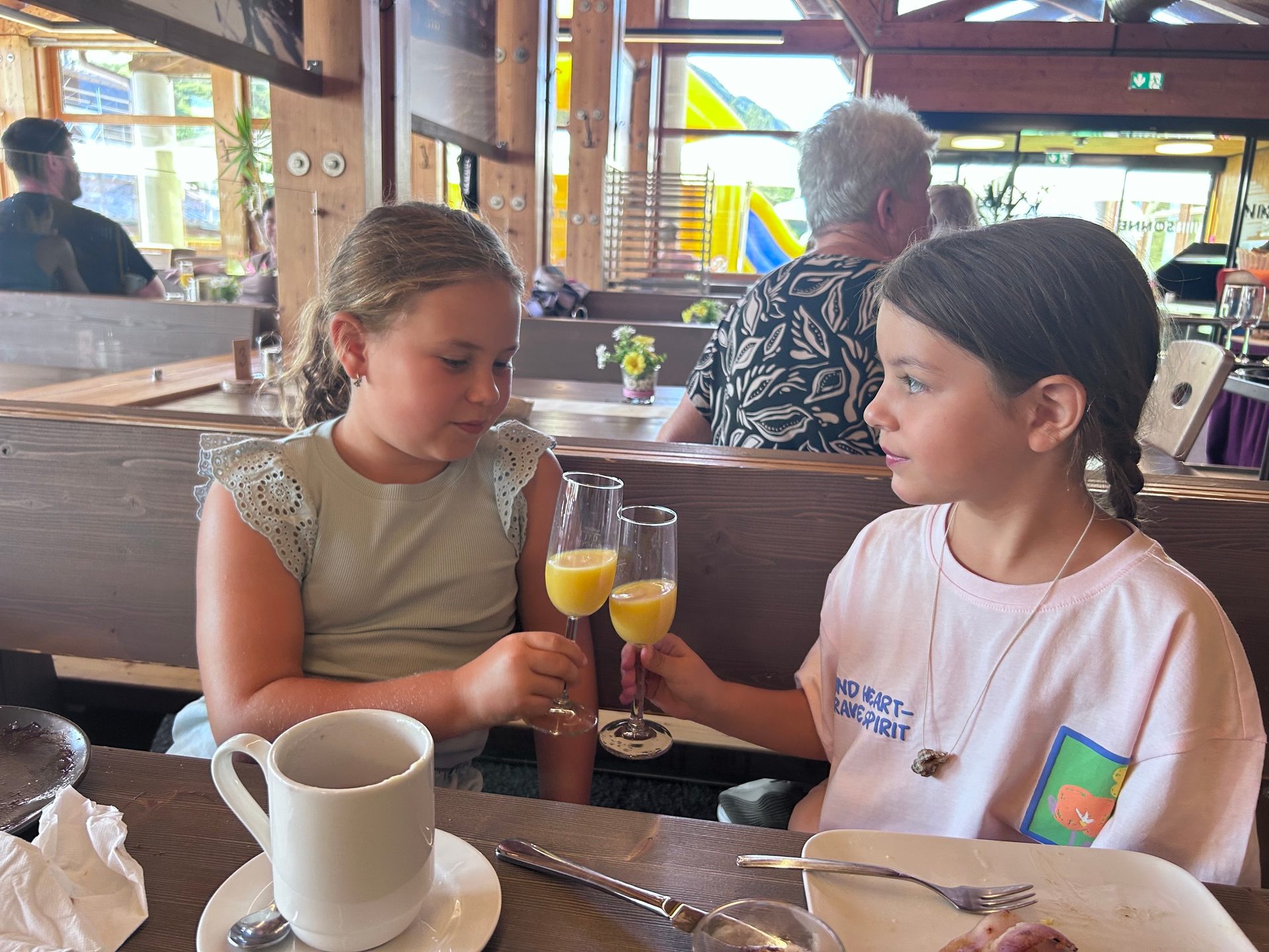 Two young girls clinking glasses filled with orange juice in a restaurant, looking at each other.